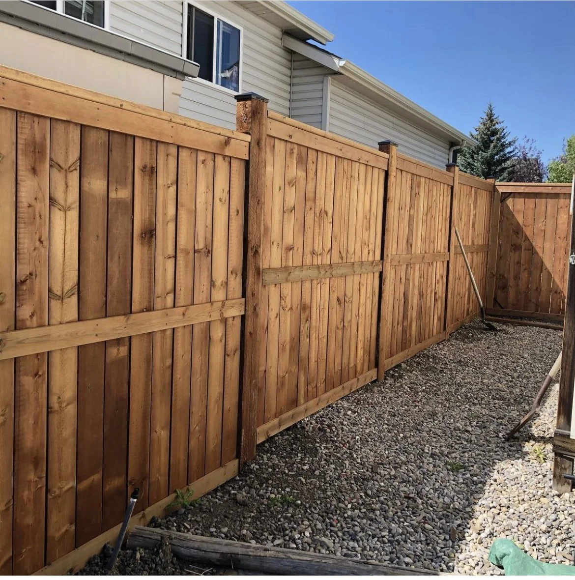 A newly built wooden privacy fence with vertical planks and horizontal support beams, running along the side of a residential yard under a clear blue sky. Part of a white house with siding and a window is visible. Gravel covers the ground, and a garden tool is leaning against the fence.