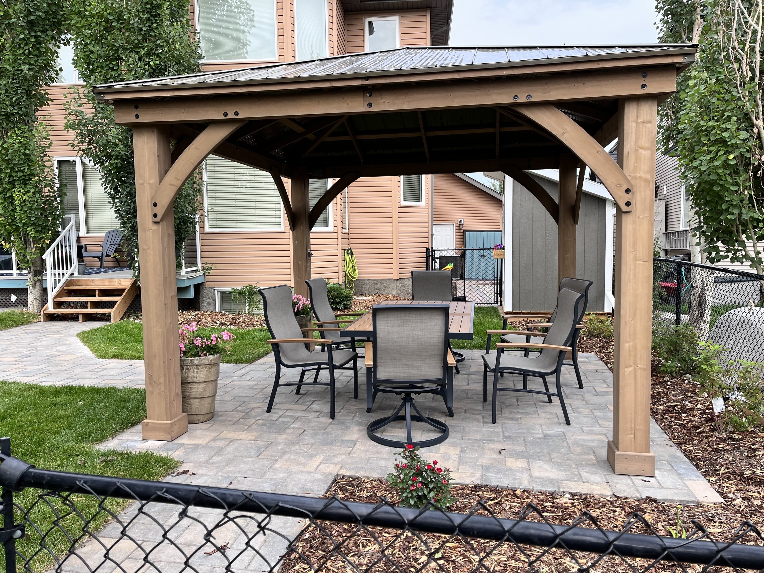 Backyard patio with a wooden gazebo, metal patio table with six chairs, potted plants, and a stone paver floor. A house with beige siding and multiple windows is in the background. Chain-link fence surrounds the yard.