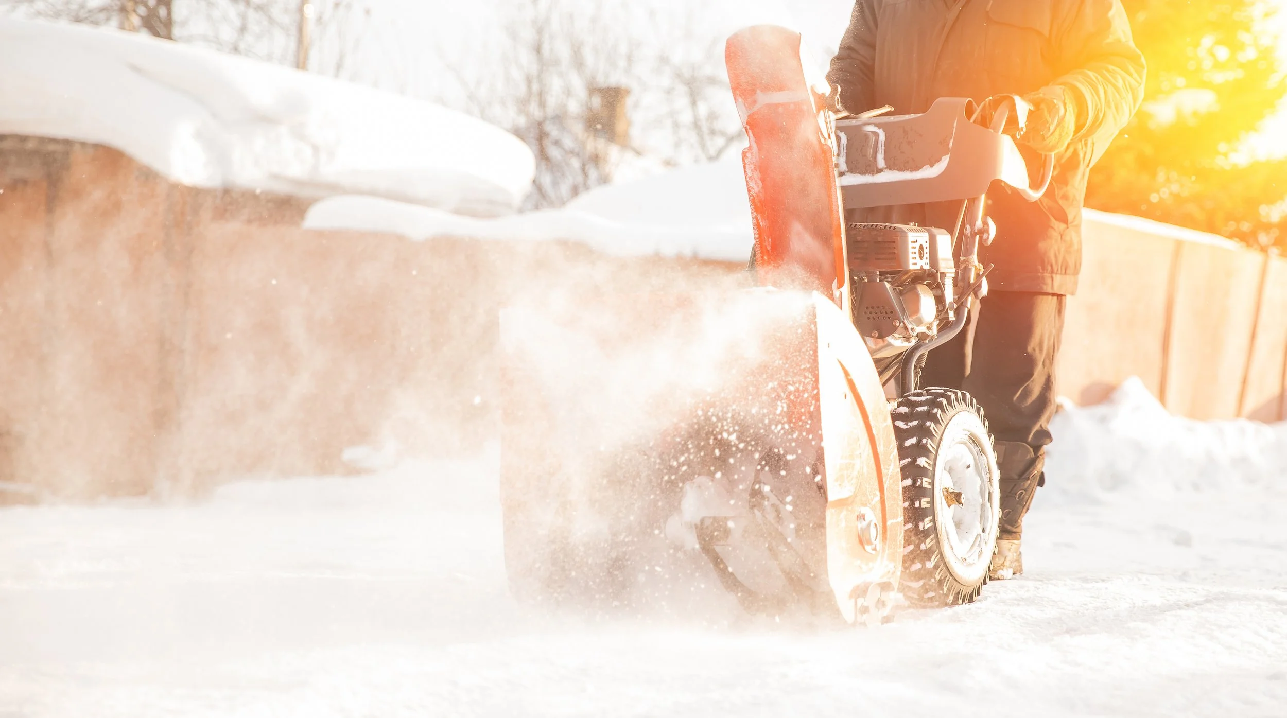 Person using a snow blower to clear snow from a driveway on a sunny winter day.