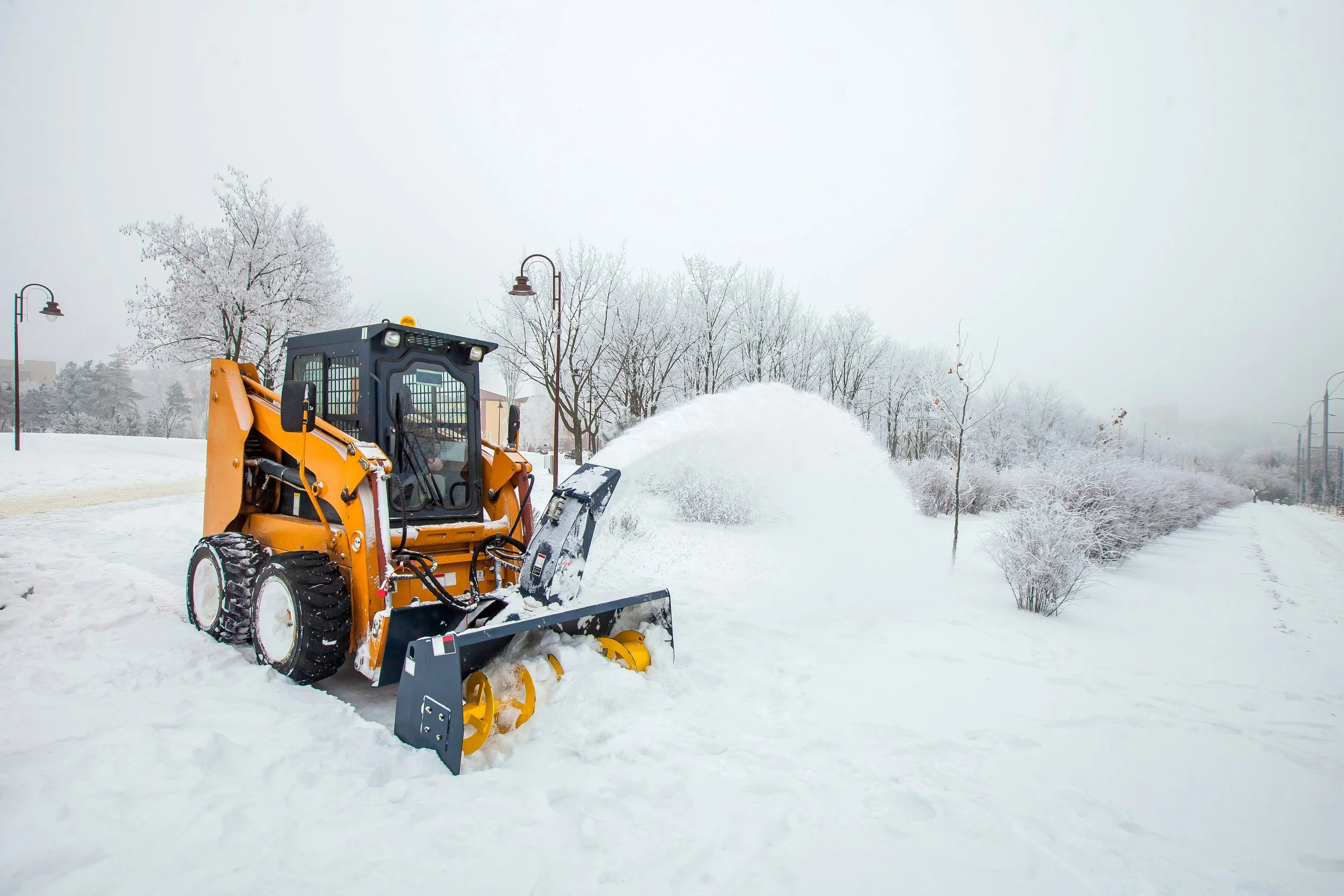 A yellow snow plow clearing snow in a park or outdoor area, with snow-covered trees and lamp posts in the background.