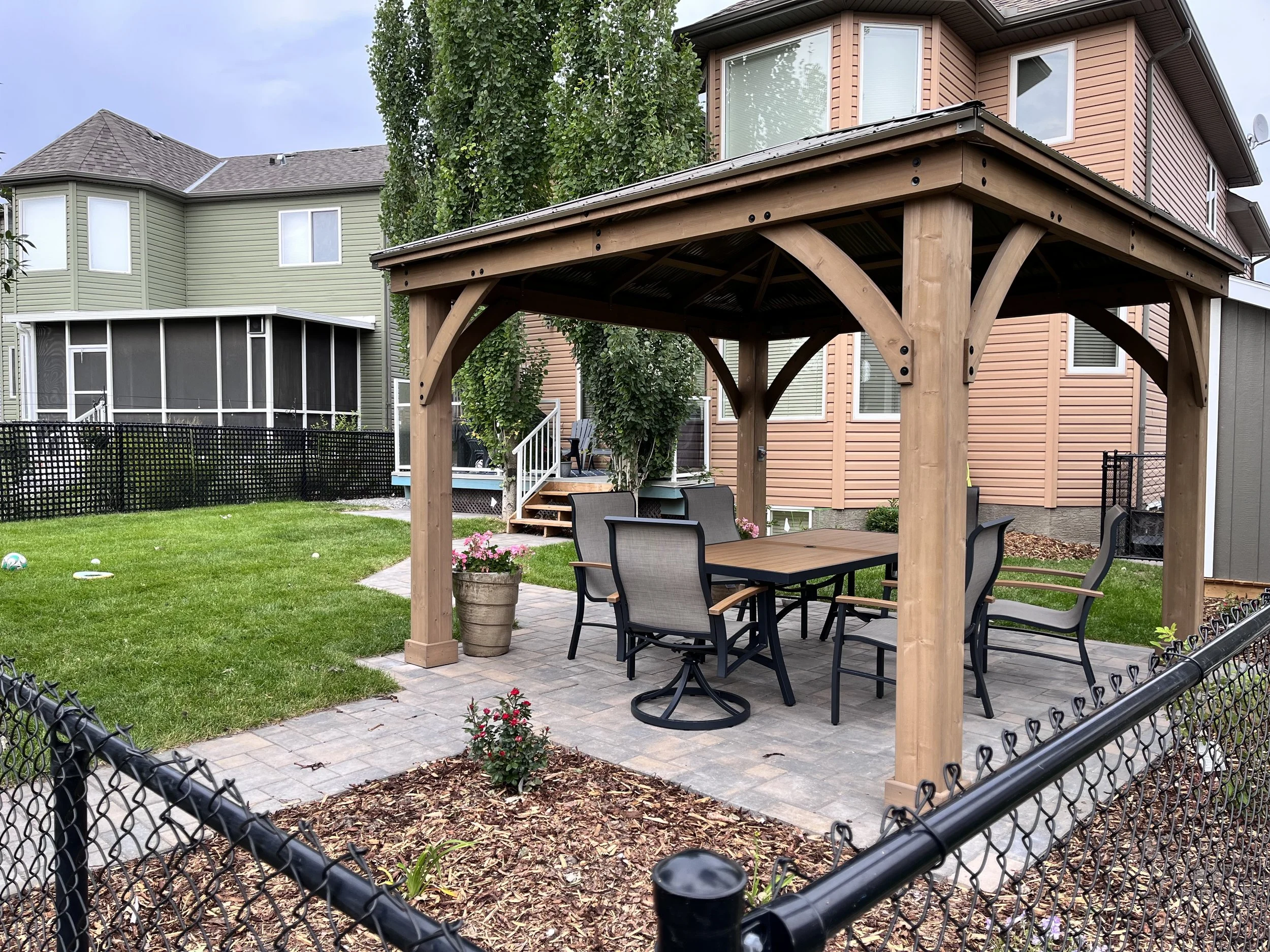 A backyard with a wooden pergola covering a patio area with outdoor chairs and a table. There are small flower pots and a flower bed with pink flowers in the foreground. The yard is enclosed with a black chain-link fence. In the background, there are neighboring houses painted in pastel colors with screened-in porches and a tree.