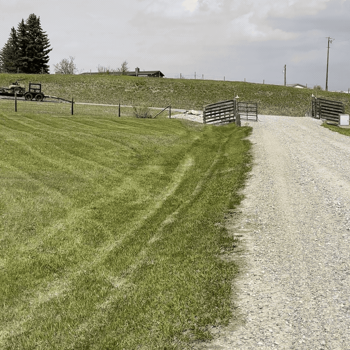 A gravel driveway leading to a farm gate with well-maintained green grass on either side and a small trailer and utility vehicle parked near a fence.
