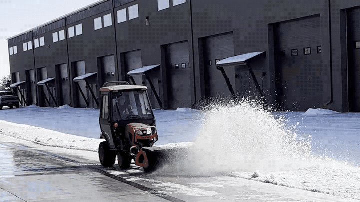 A small snow plow clearing snow from a driveway in front of a building with loading docks in an industrial area.