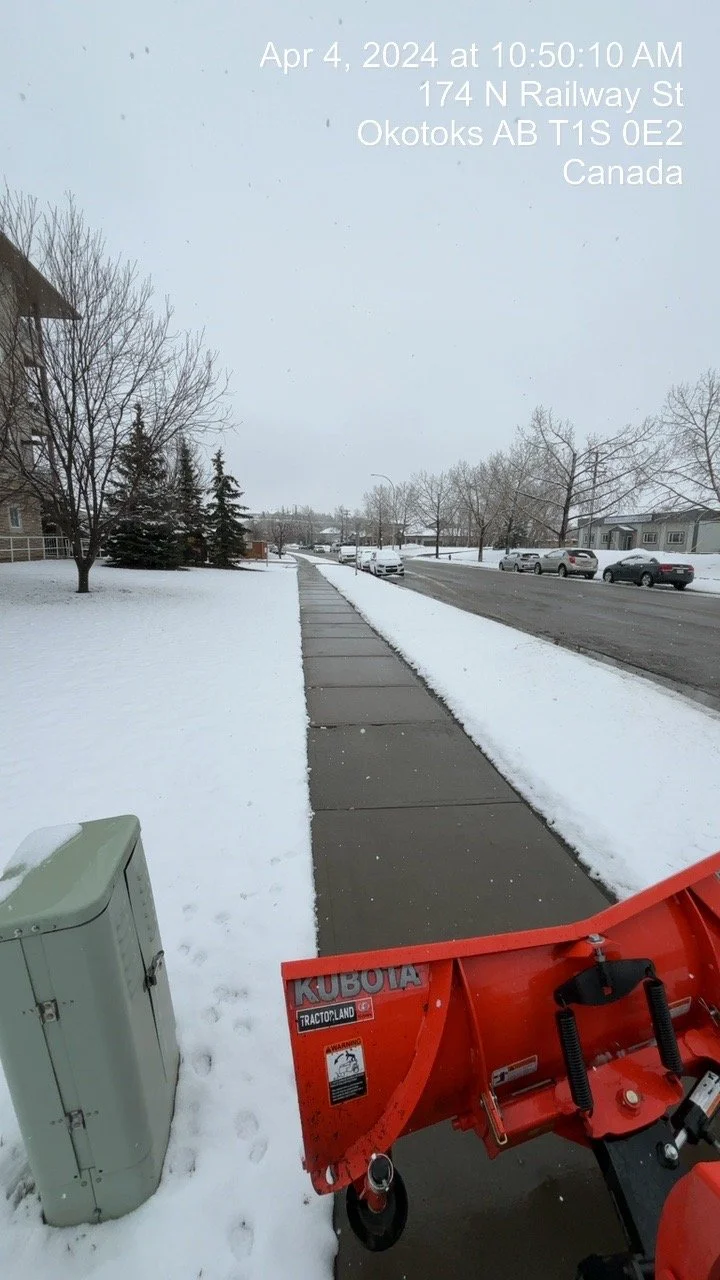 Snowy sidewalk and street with parked cars and leafless trees, with a snow removal machine in the foreground near a utility box, and a timestamp overlay at the top.