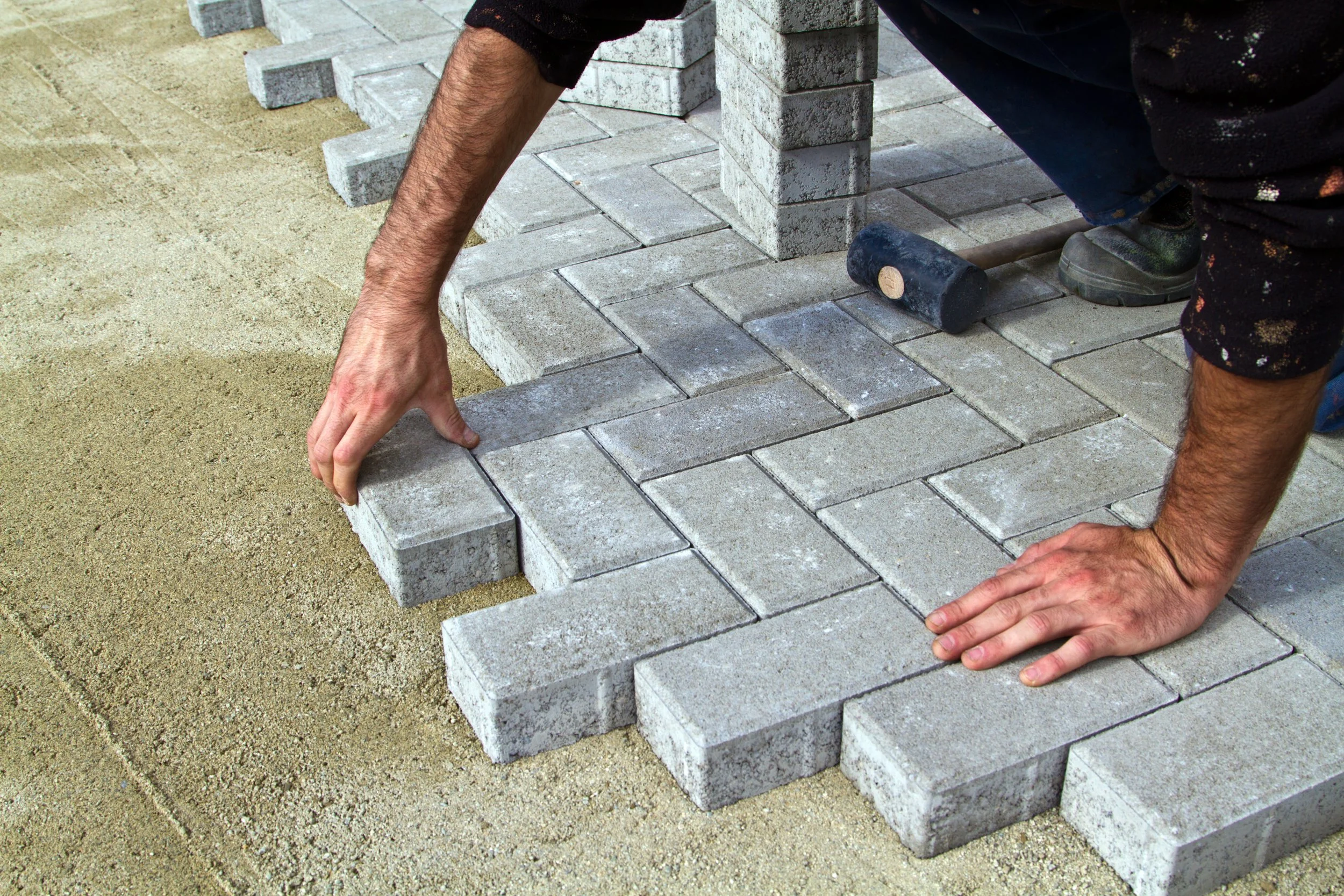 Person laying down bricks for a pathway. They are on their hands and knees, placing bricks on a sandy surface. The bricks are grey and rectangular. The person is wearing dark clothing and sneakers.