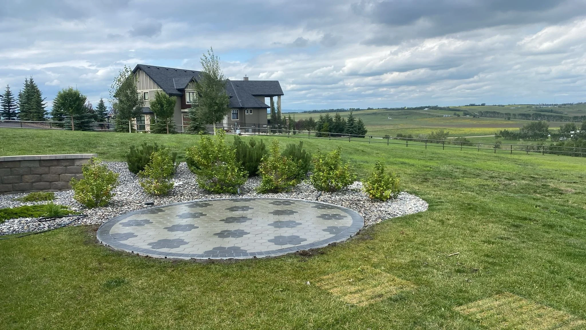 A yard with a circular paving stone patio, rocks, bushes, and grass, overlooking a house with a black roof and a fenced yard in a rural area with rolling hills and cloudy sky.