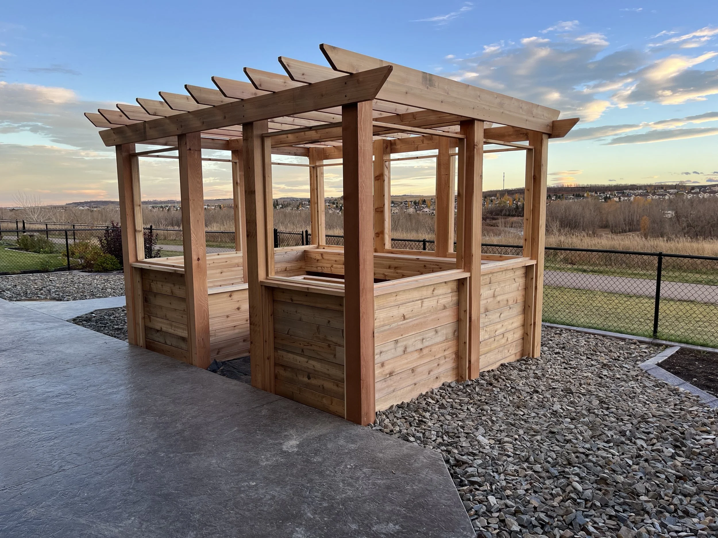 A wooden pergola structure with a partially open roof viewed outdoors during sunset, with a scenic background of a neighborhood and open land.