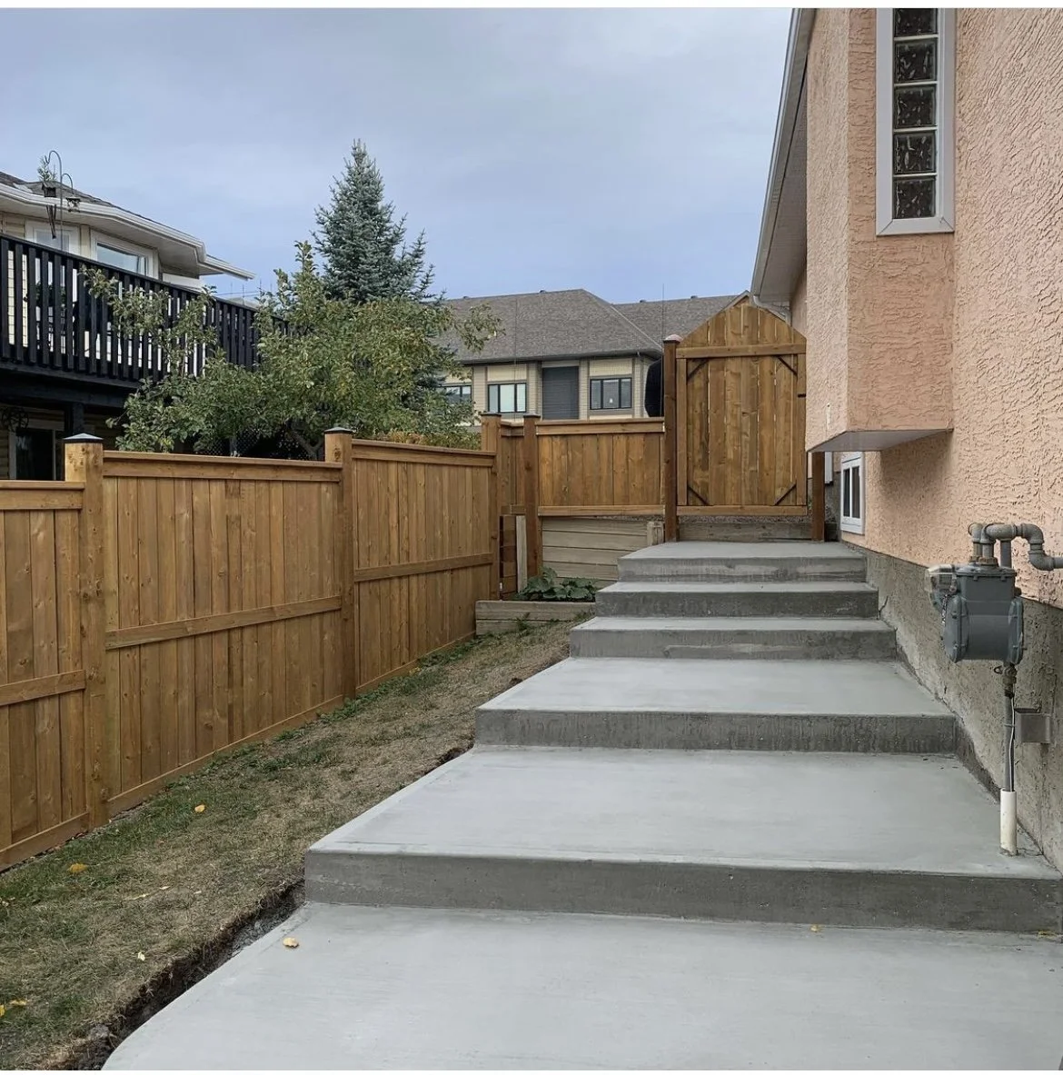 Newly constructed concrete stairs leading up to a wooden gate and fence behind a house with textured peach-colored wall, with neighboring houses visible in the background.