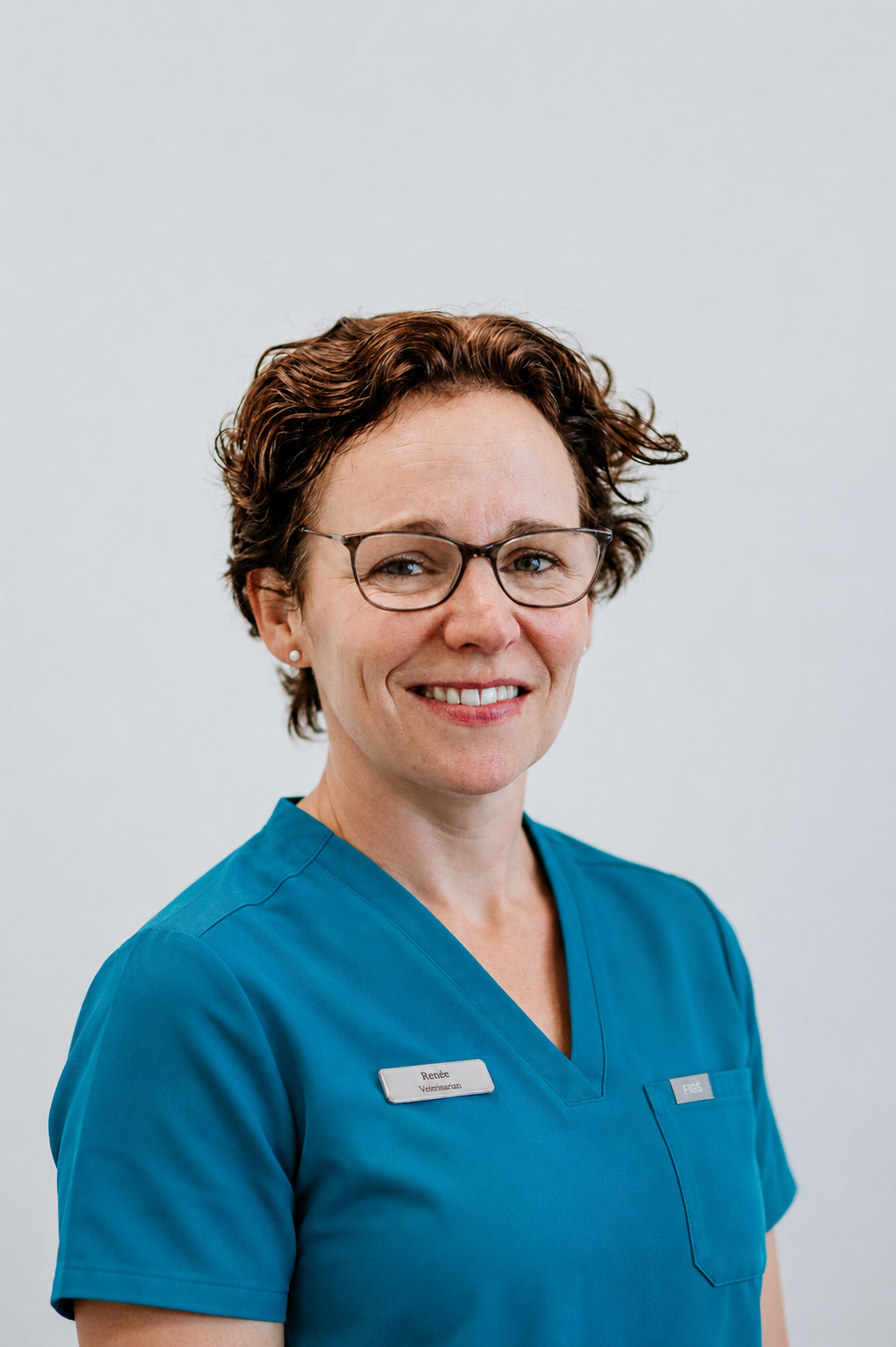 A veterinarian with short, curly brown hair and glasses, wearing a blue medical uniform with a badge, smiling against a plain white background.