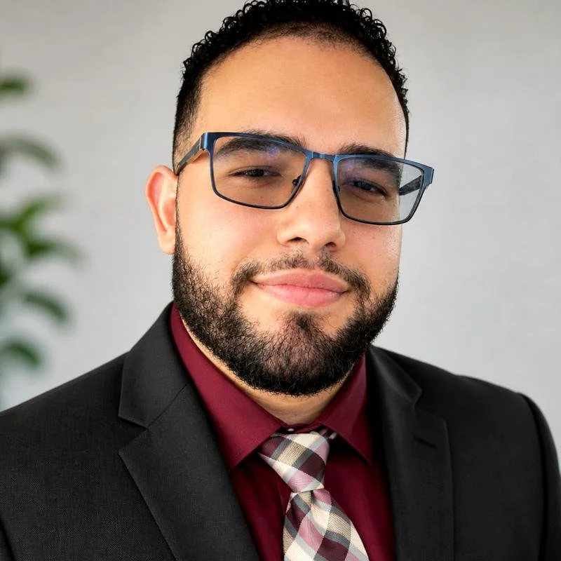 A man with a beard and glasses wearing a black suit, maroon shirt, and plaid tie, smiling in a professional setting.