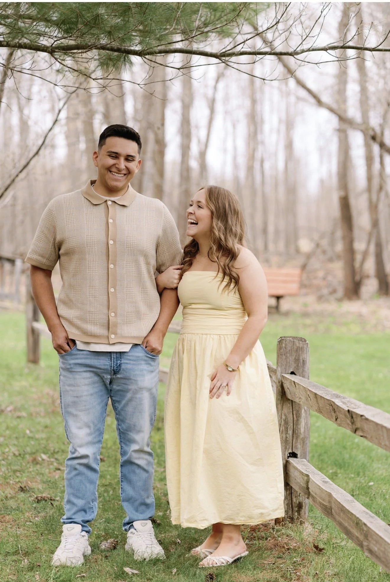 A smiling young man and woman standing outdoors in a park, surrounded by leafless trees, near a wooden fence, during spring or fall.