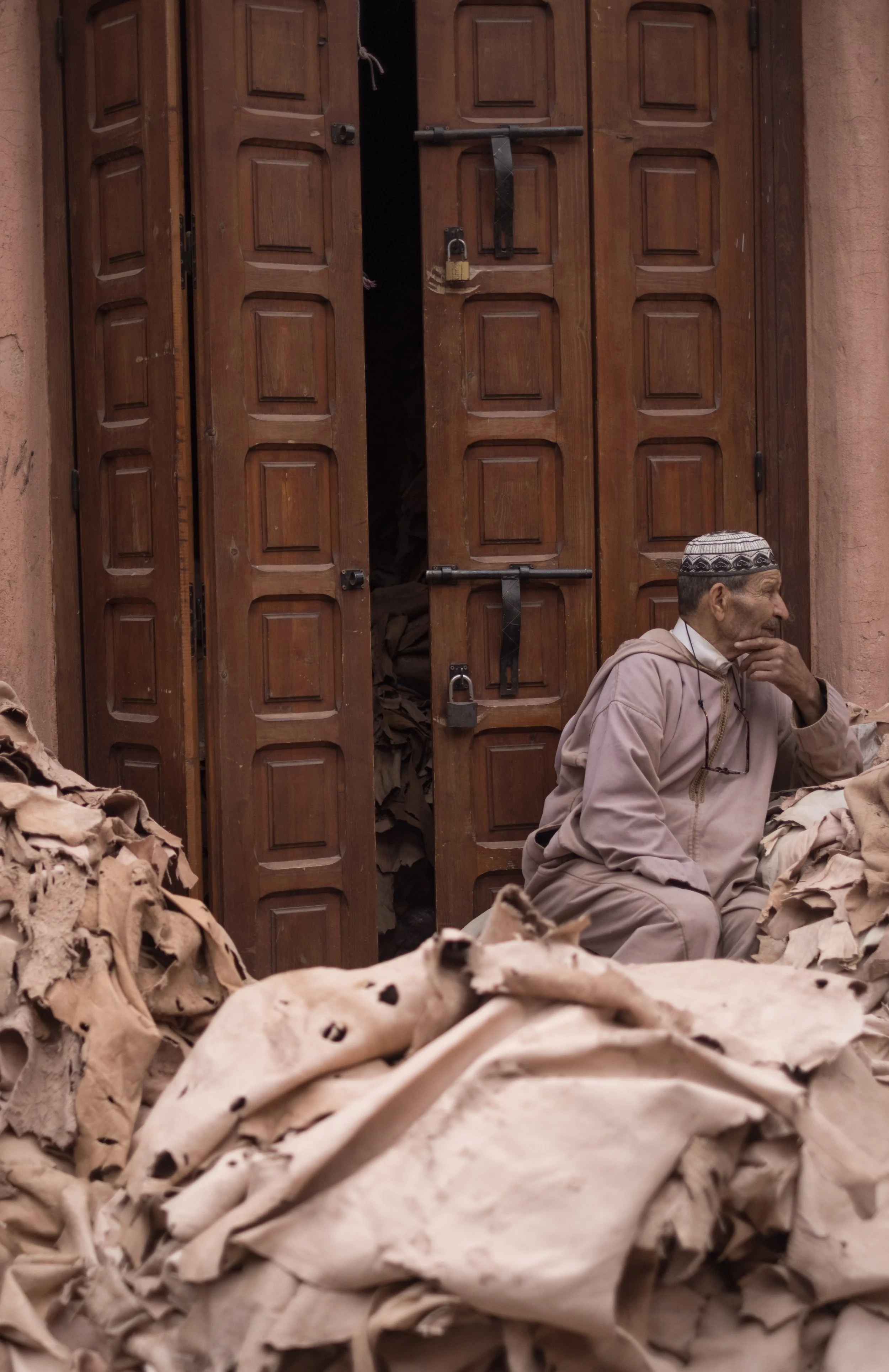 An elderly man wearing a traditional cap sitting on the ground, surrounded by piles of dried plant material, in front of a wooden door with padlocks.