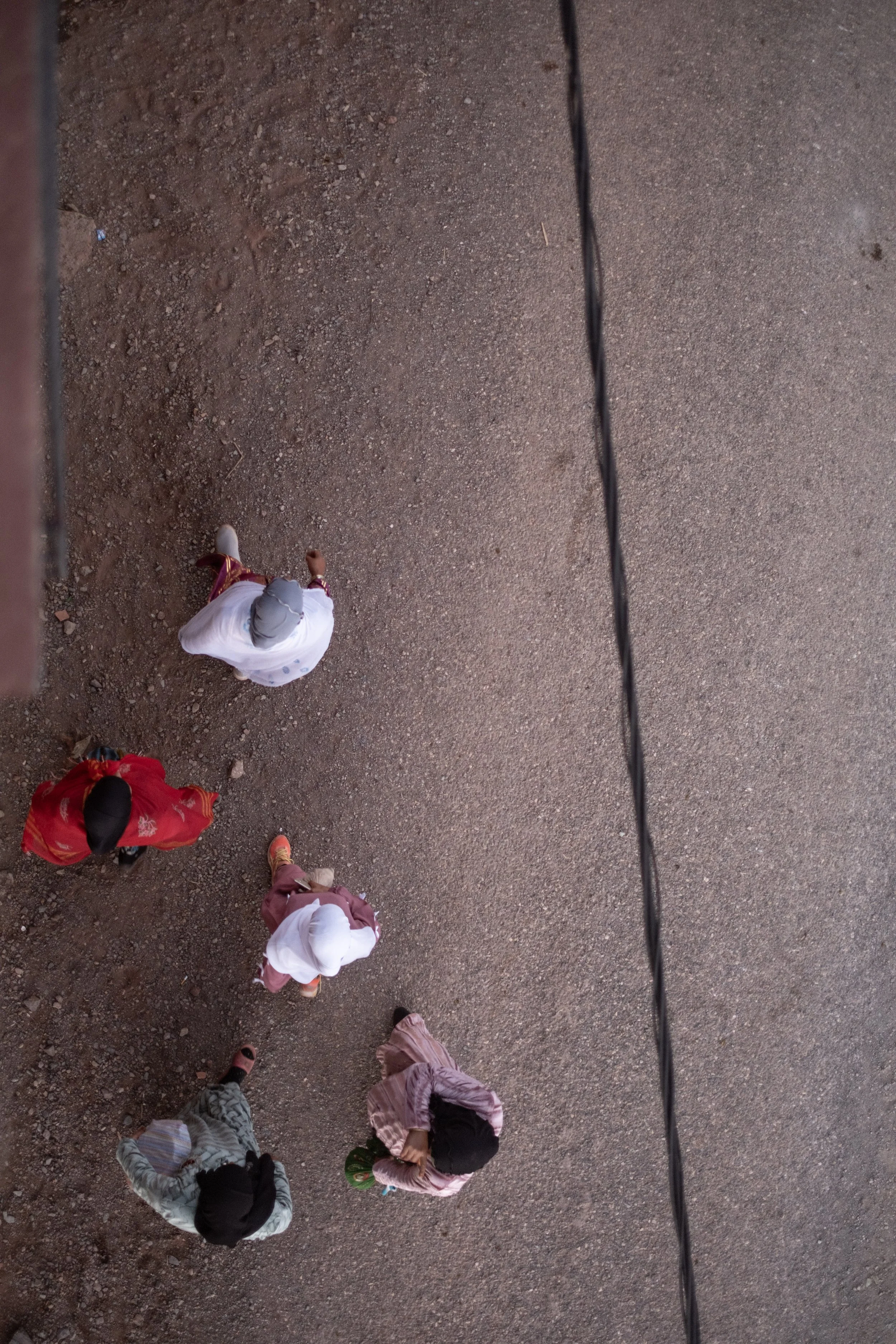 From a high angle, people are walking on a dirt ground, some wearing hijabs and colorful clothing, with a thin black cable running vertically through the right side of the image.