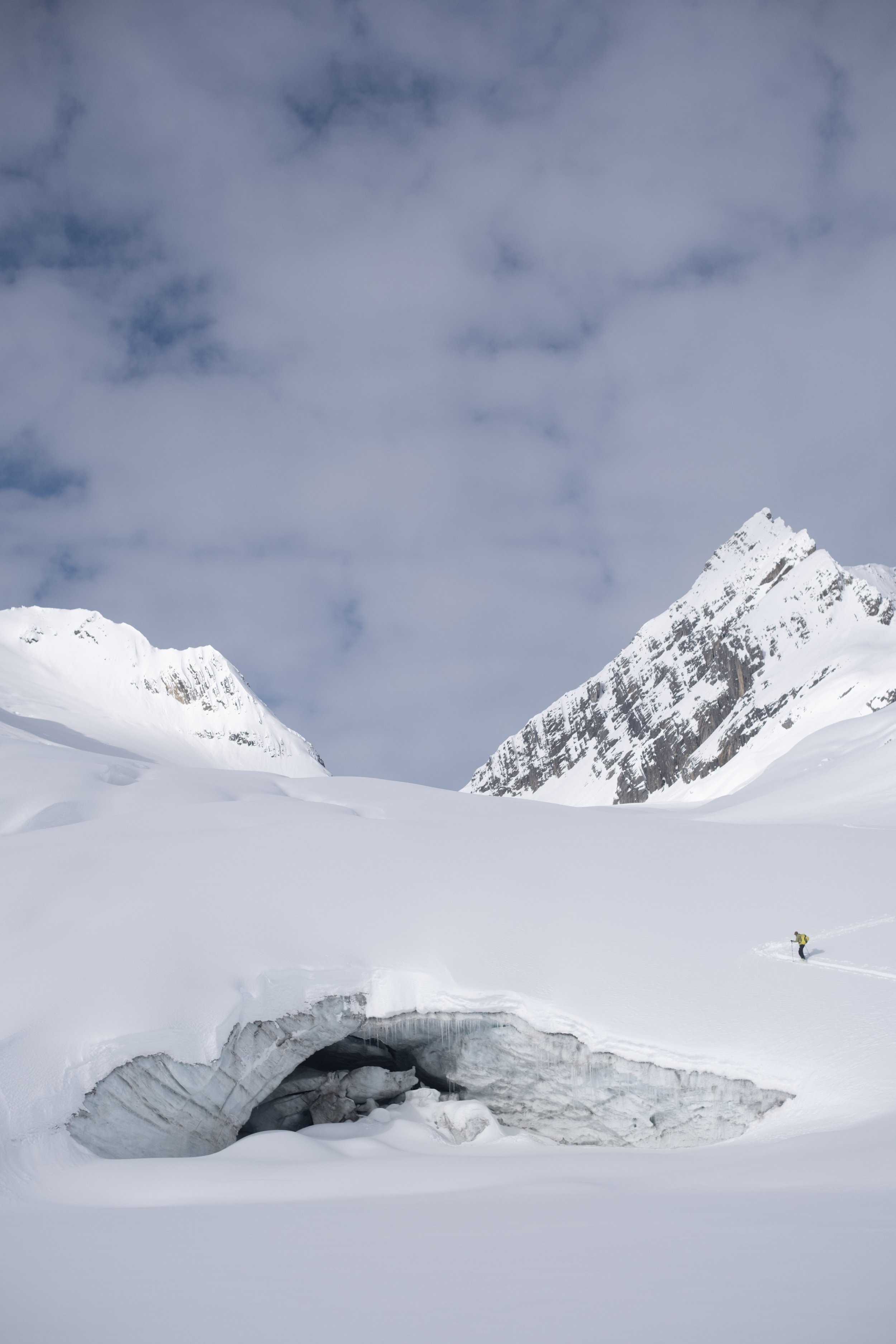 Snow-covered mountains with an icy crevasse in the foreground and a climber in yellow gear on the right side.