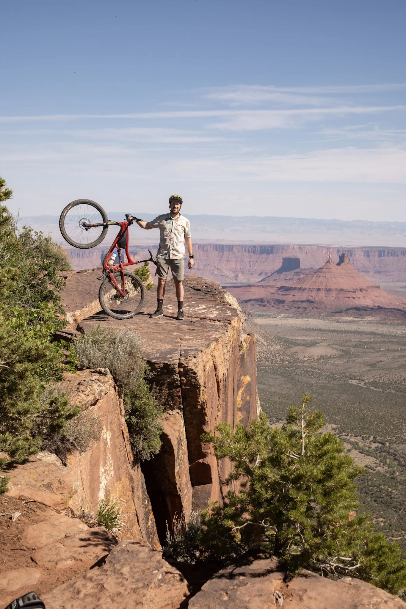 A man in outdoor clothing and a helmet holding a red bicycle on a cliff edge overlooking a desert canyon landscape with rock formations and sparse vegetation under a blue sky.