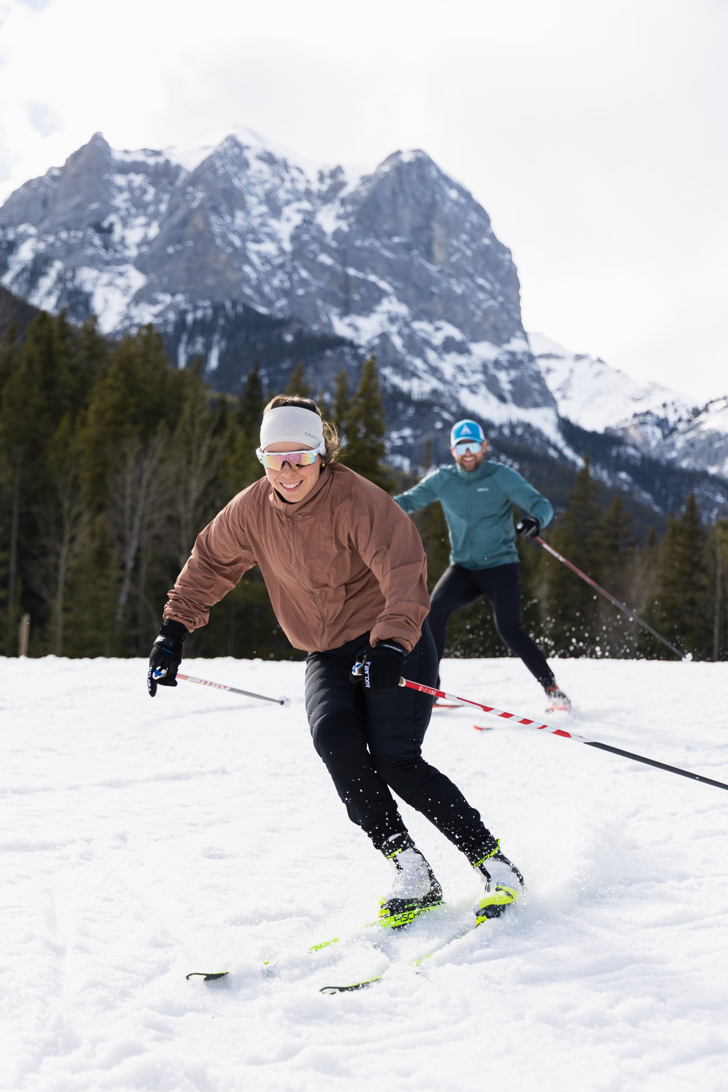Two people are skiing on snow-covered ground outdoors, with a mountain range with patches of snow and evergreen trees in the background. The woman in the foreground is smiling, wearing a brown jacket, white headband, and sunglasses. The man behind he