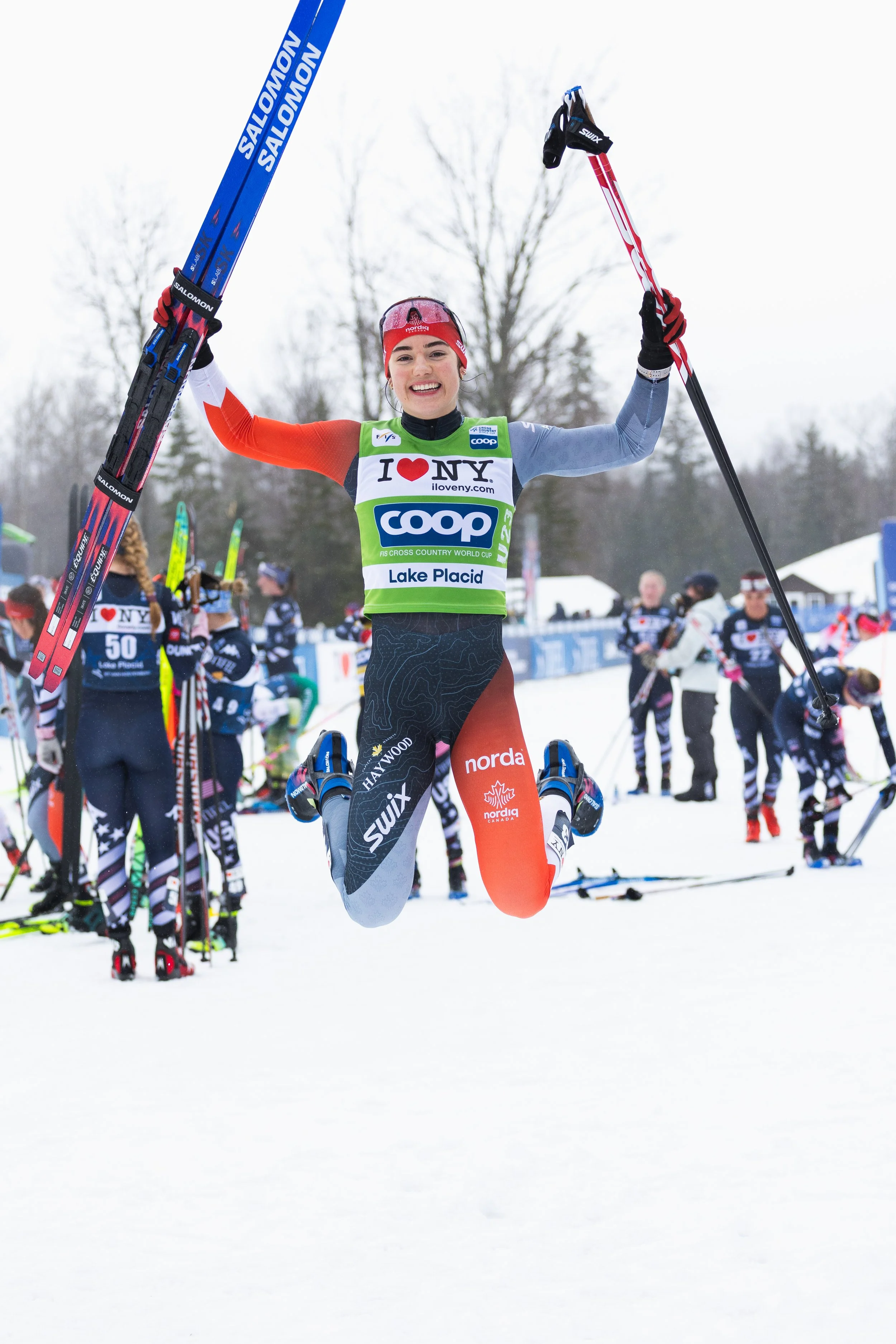 A female cross-country skier jumping in the air with skis and poles raised, smiling at a snow-covered outdoor event with other participants and spectators in the background.