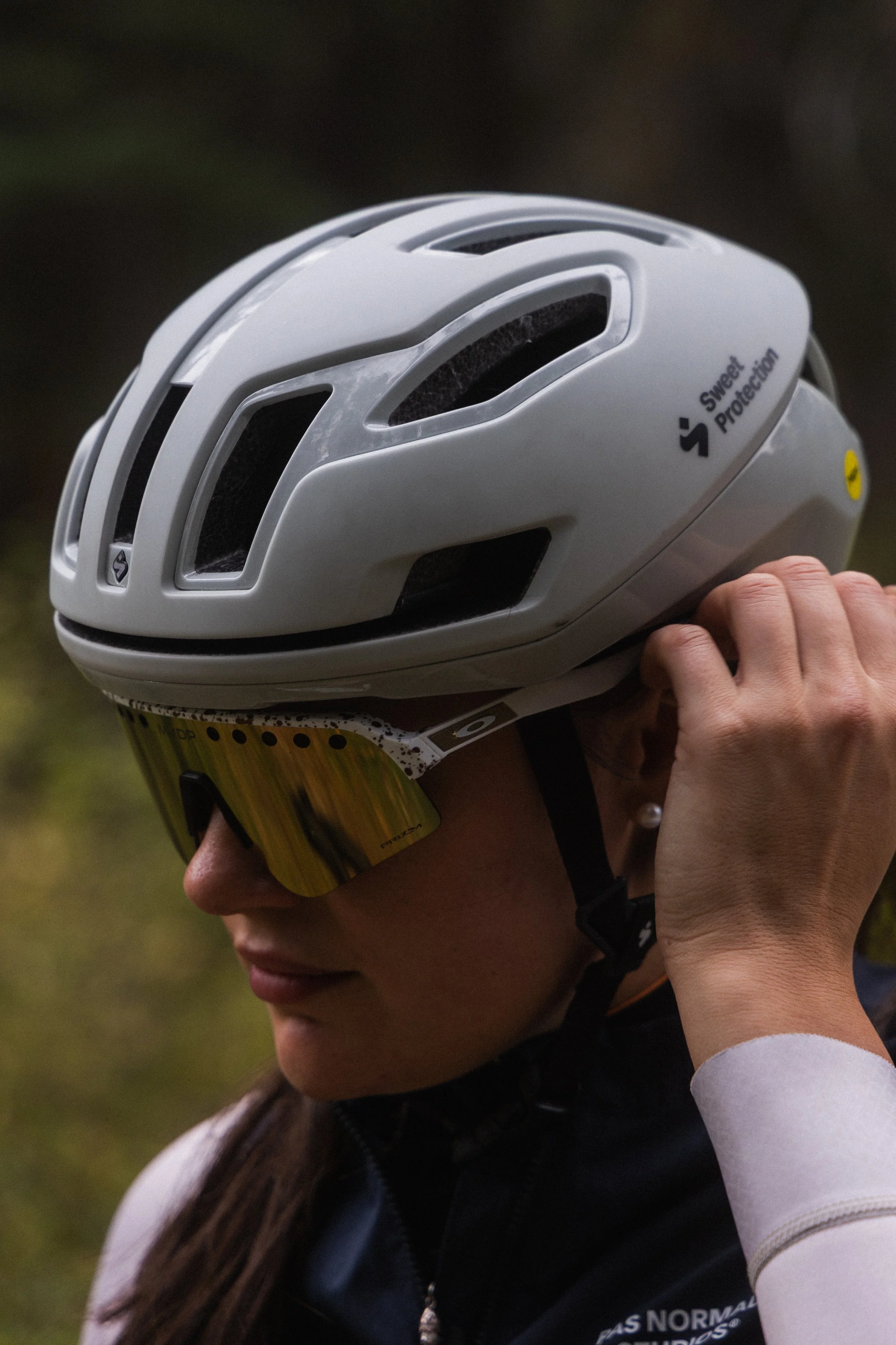 Close-up of a person wearing a white protective helmet and sunglasses, with their face partially visible. The helmet has ventilation openings and a label with the words 'Sweet Protection.'