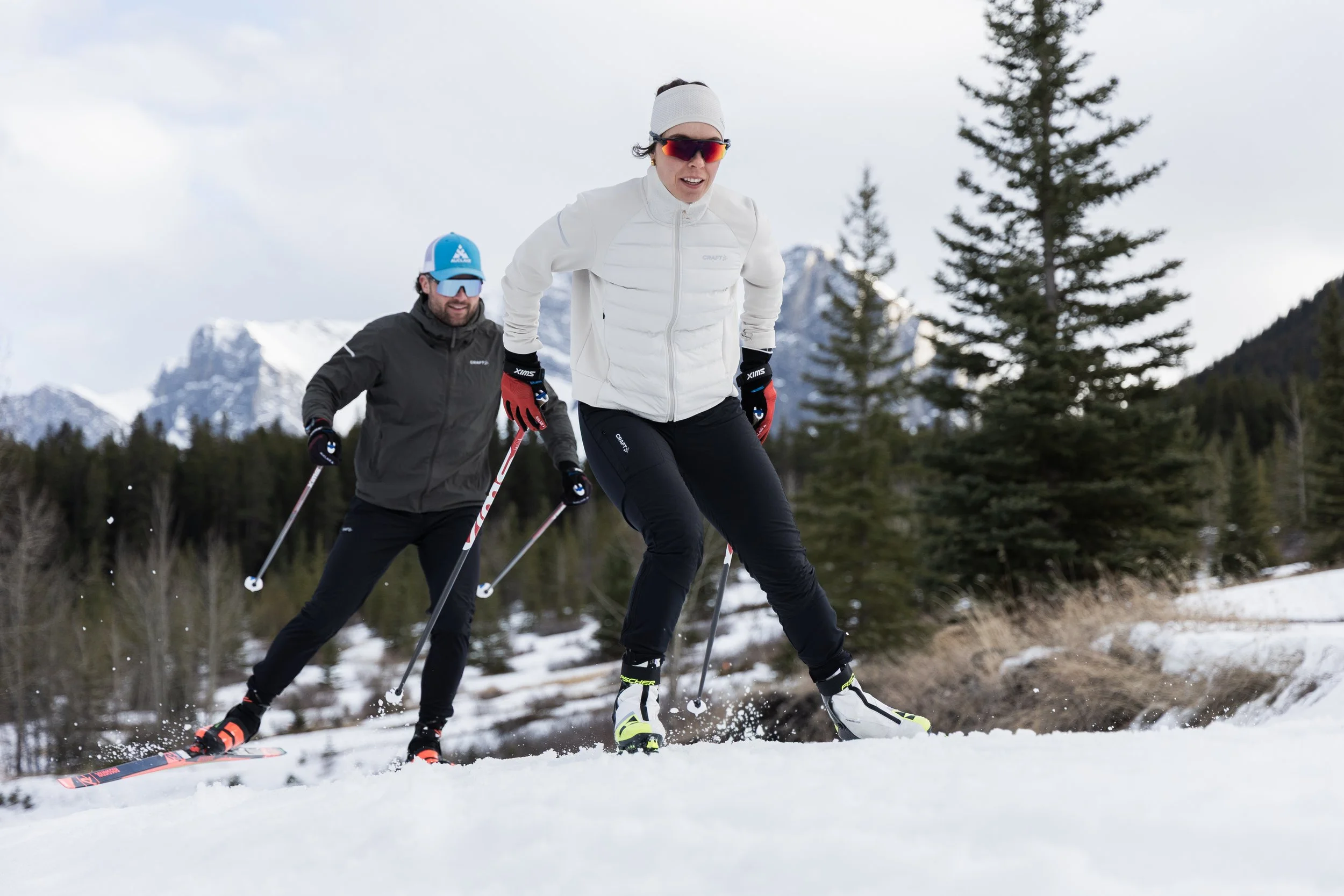 Two people cross-country skiing in a snowy landscape with mountains and trees in the background. They are dressed in winter sports gear, including jackets, gloves, and sunglasses.