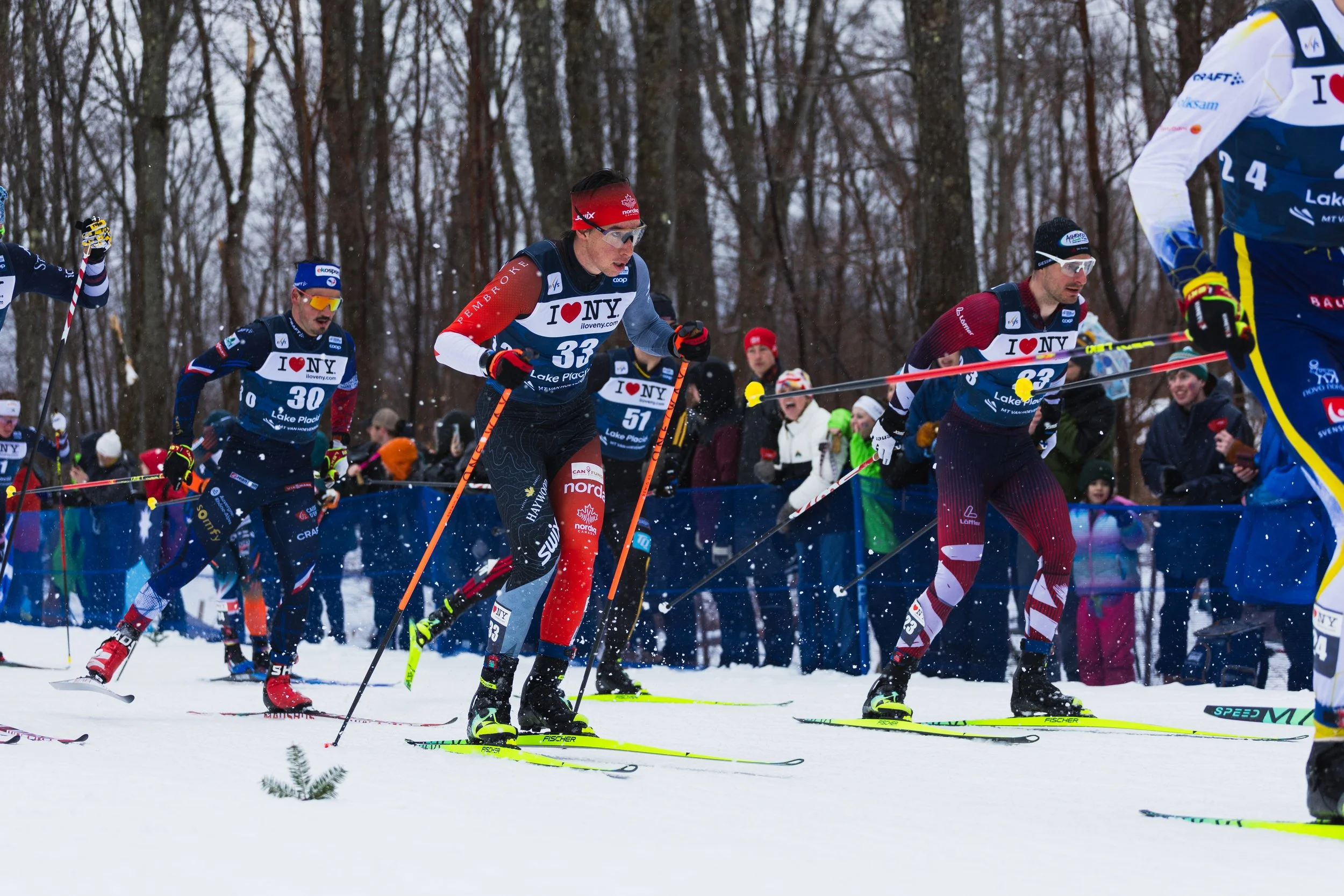 Cross-country skiers racing outdoors in a snow-covered forest with spectators watching, some taking photos, during a competitive event.