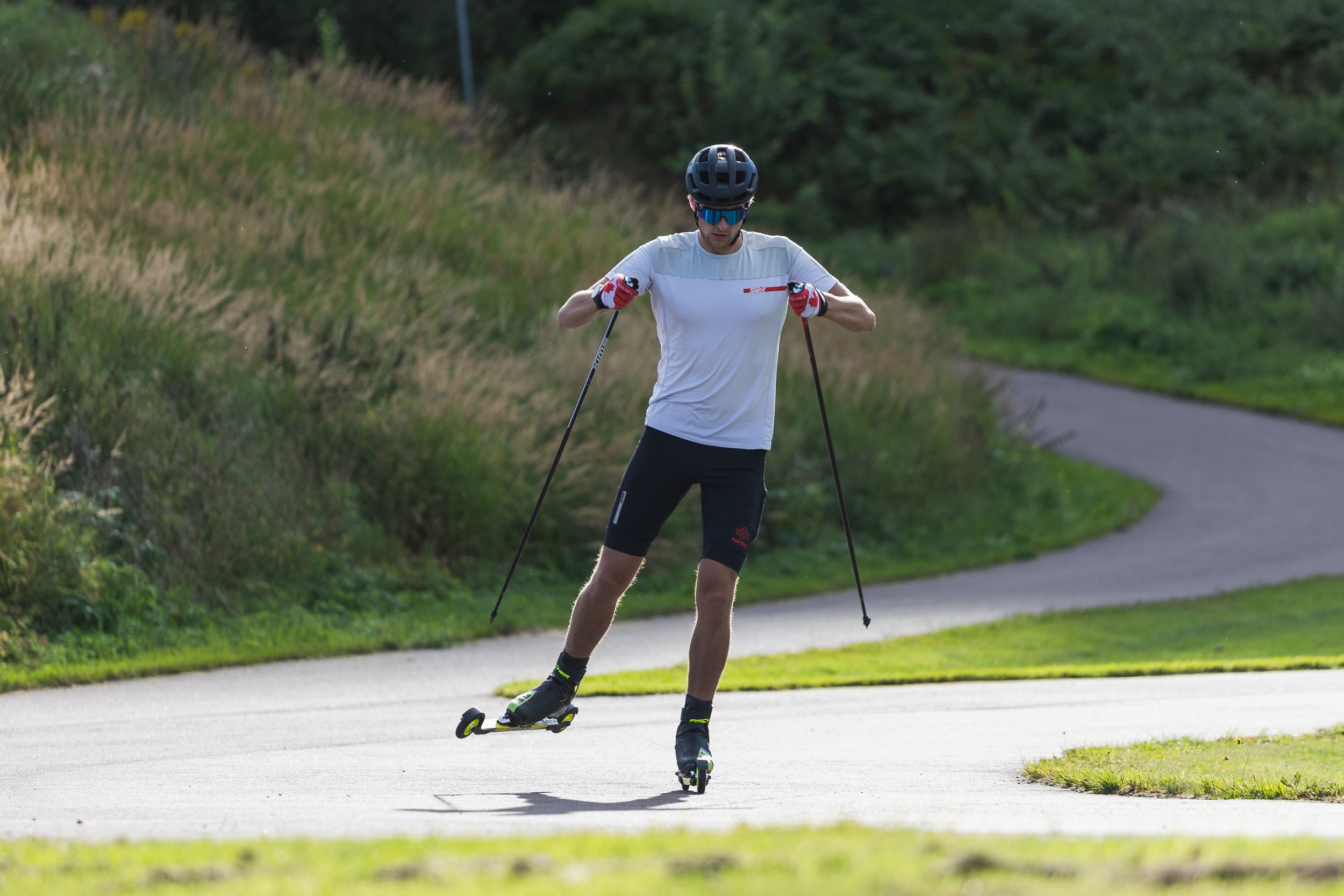 A man wearing a helmet, sunglasses, and athletic clothing is skateboarding on a paved path through a green park.