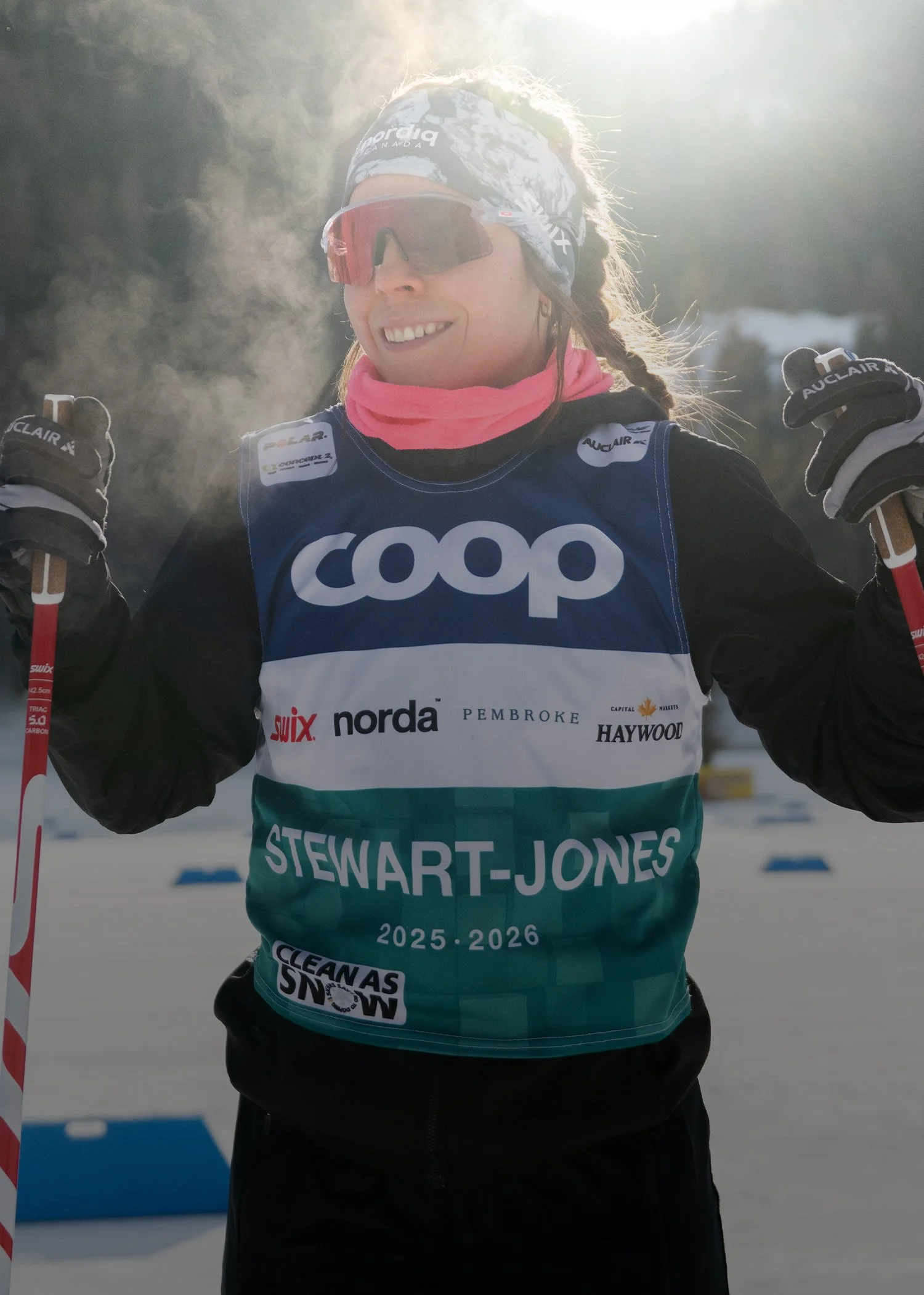 Female cross-country skier in a racing suit and headband, holding ski poles, smiling outdoors in winter sunlight with snow in the background.