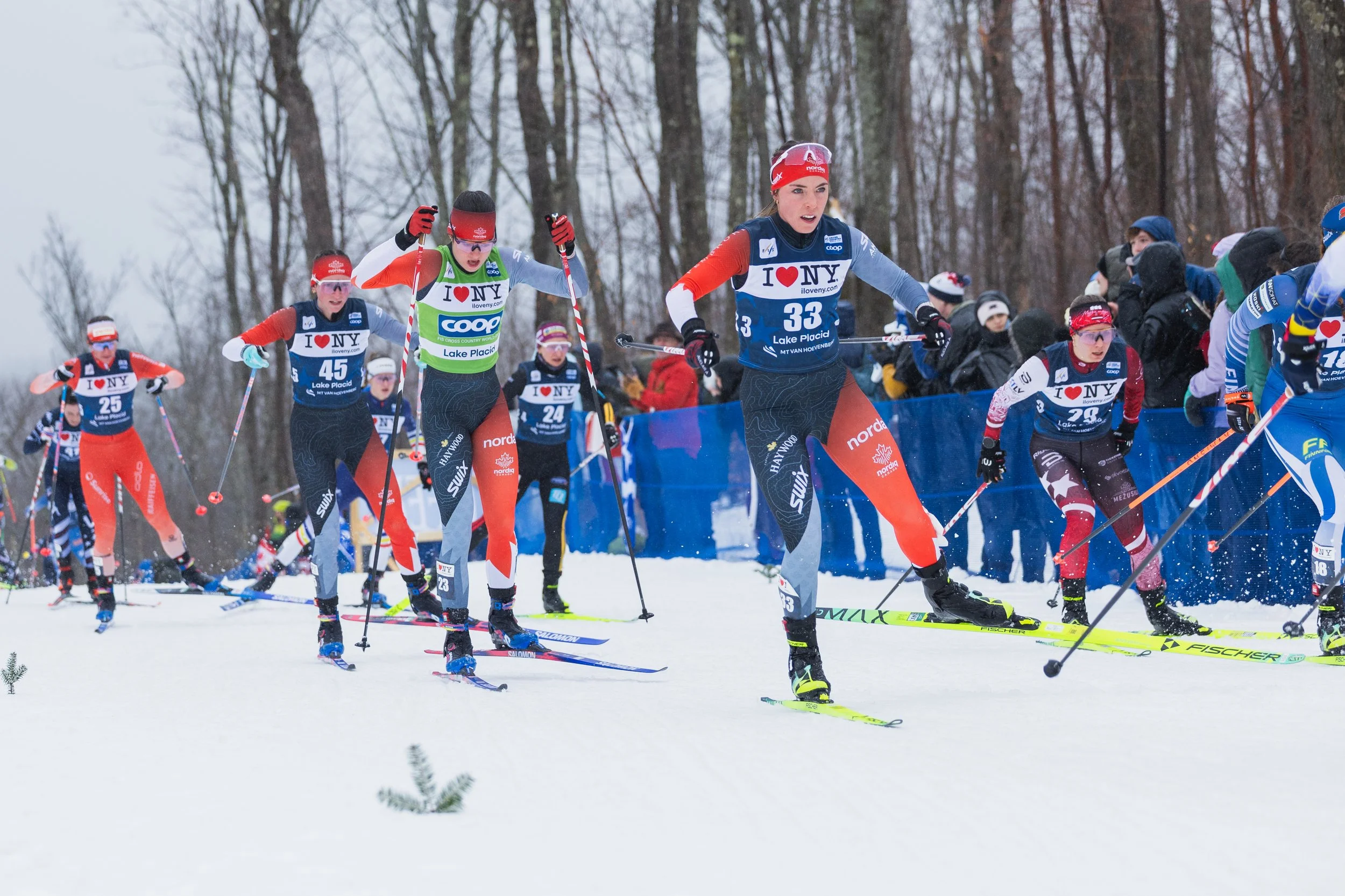 A group of female cross-country skiers competing in a race in a snowy forest, wearing racing bibs with the logo 'I ❤️ NY' and various sponsor logos, using skis and poles, with spectators in winter clothing watching from the side.
