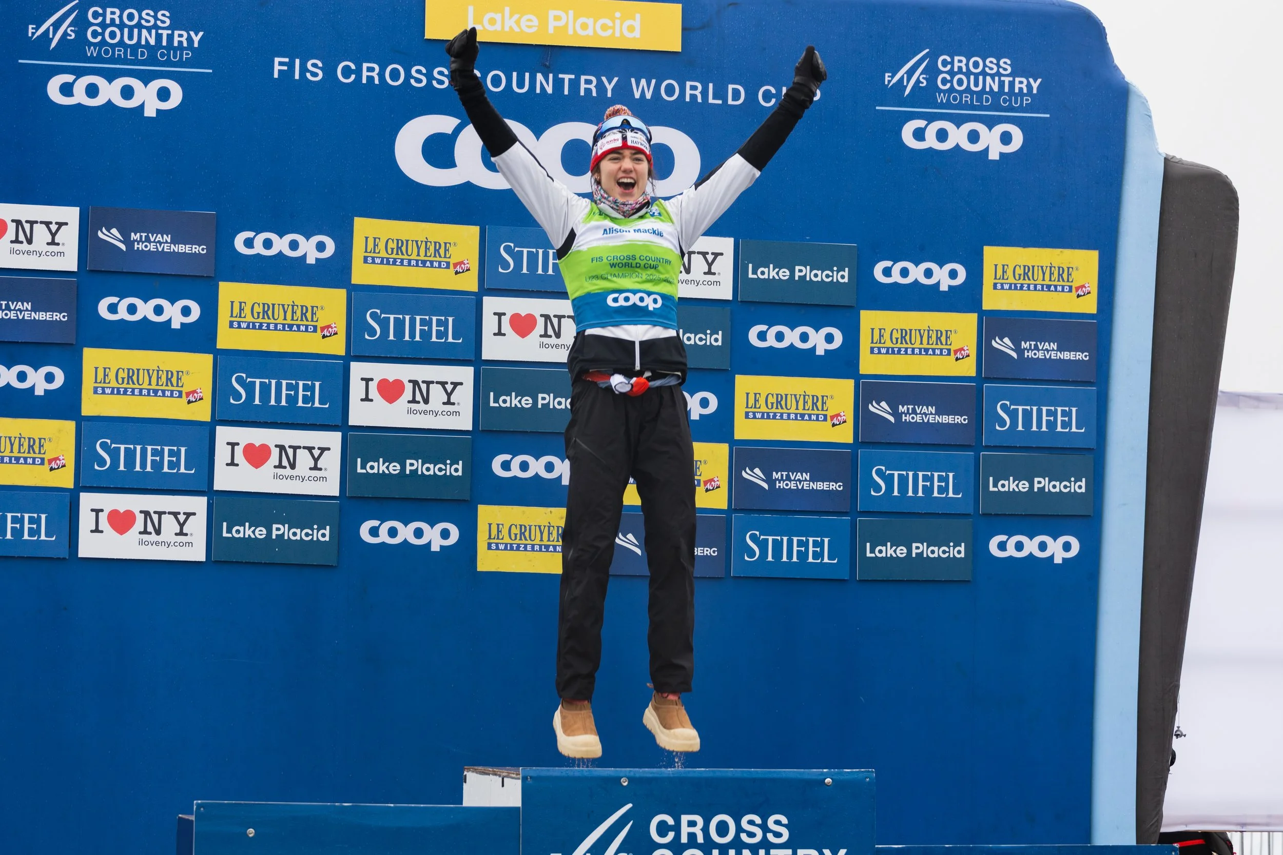 A female athlete celebrating on the podium after a cross-country skiing event at Lake Placid. She is smiling, has her arms raised, and is wearing a white and black outfit with a red, white, and blue hat. The background features a blue wall with logos