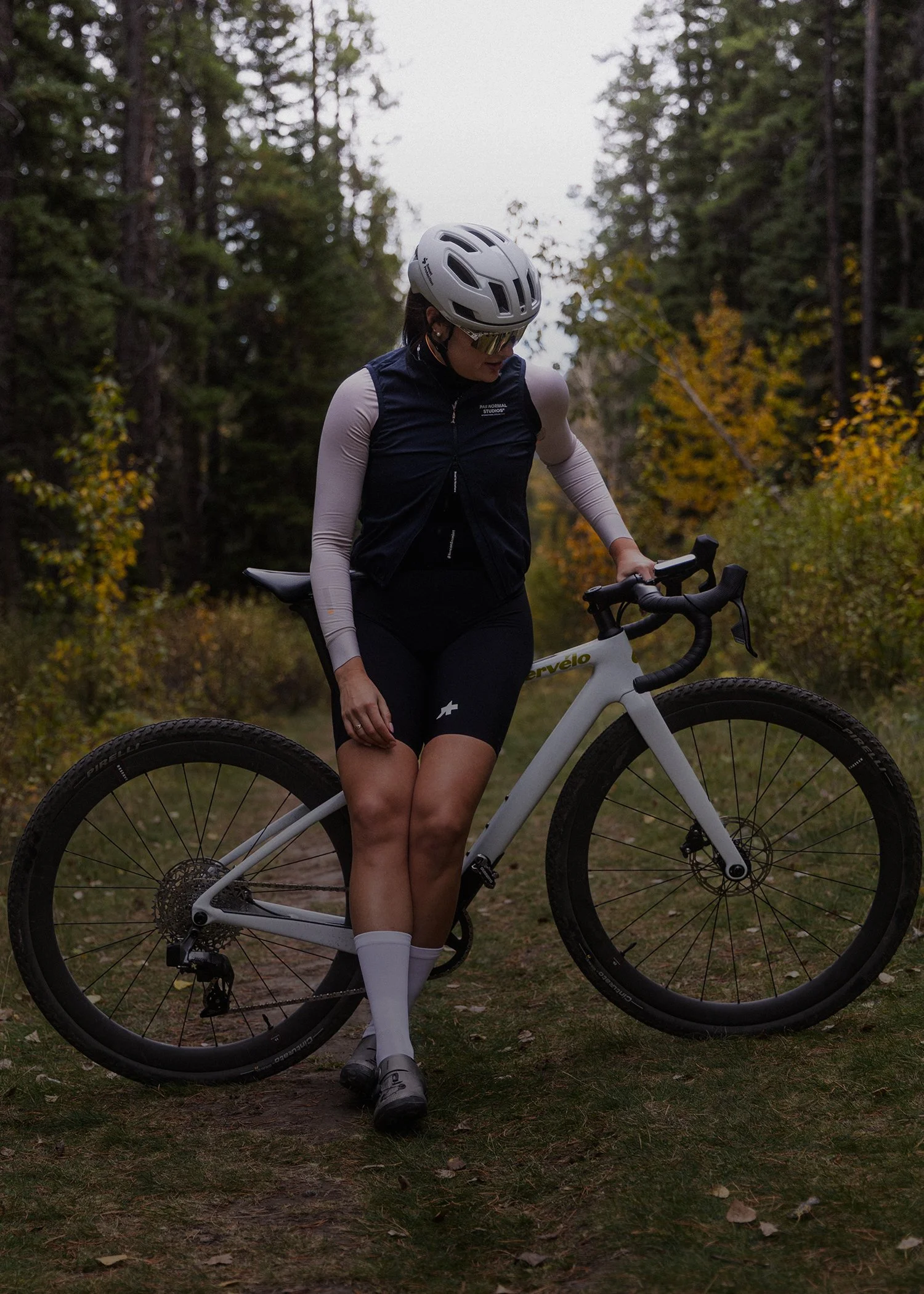 A woman wearing a white cycling helmet, sunglasses, and a black and white cycling outfit, standing with a white mountain bike on a wooded trail with autumn foliage.