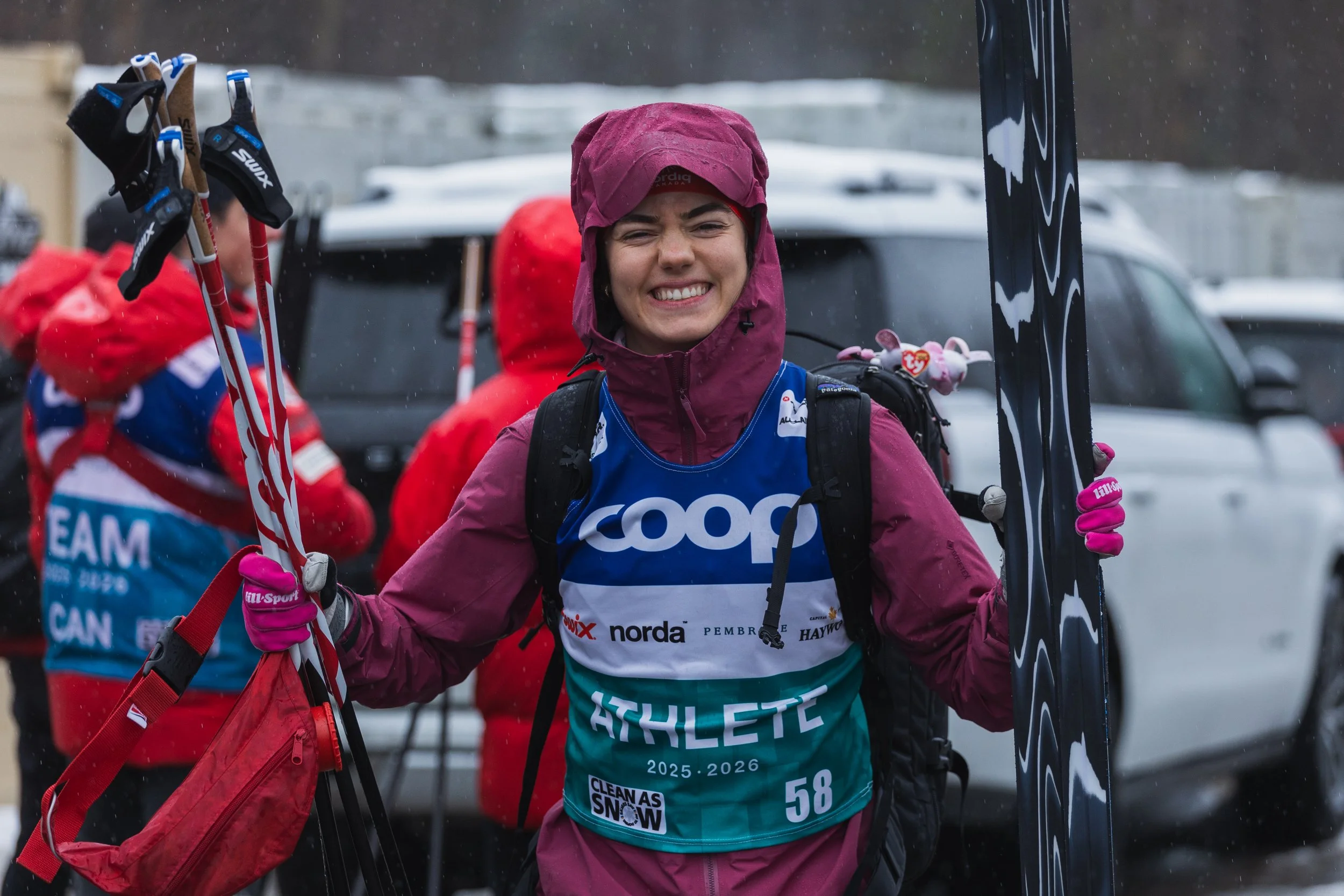 Smiling female athlete in purple rain jacket holding skis, participating in a winter race, with other racers and parked cars in the background.