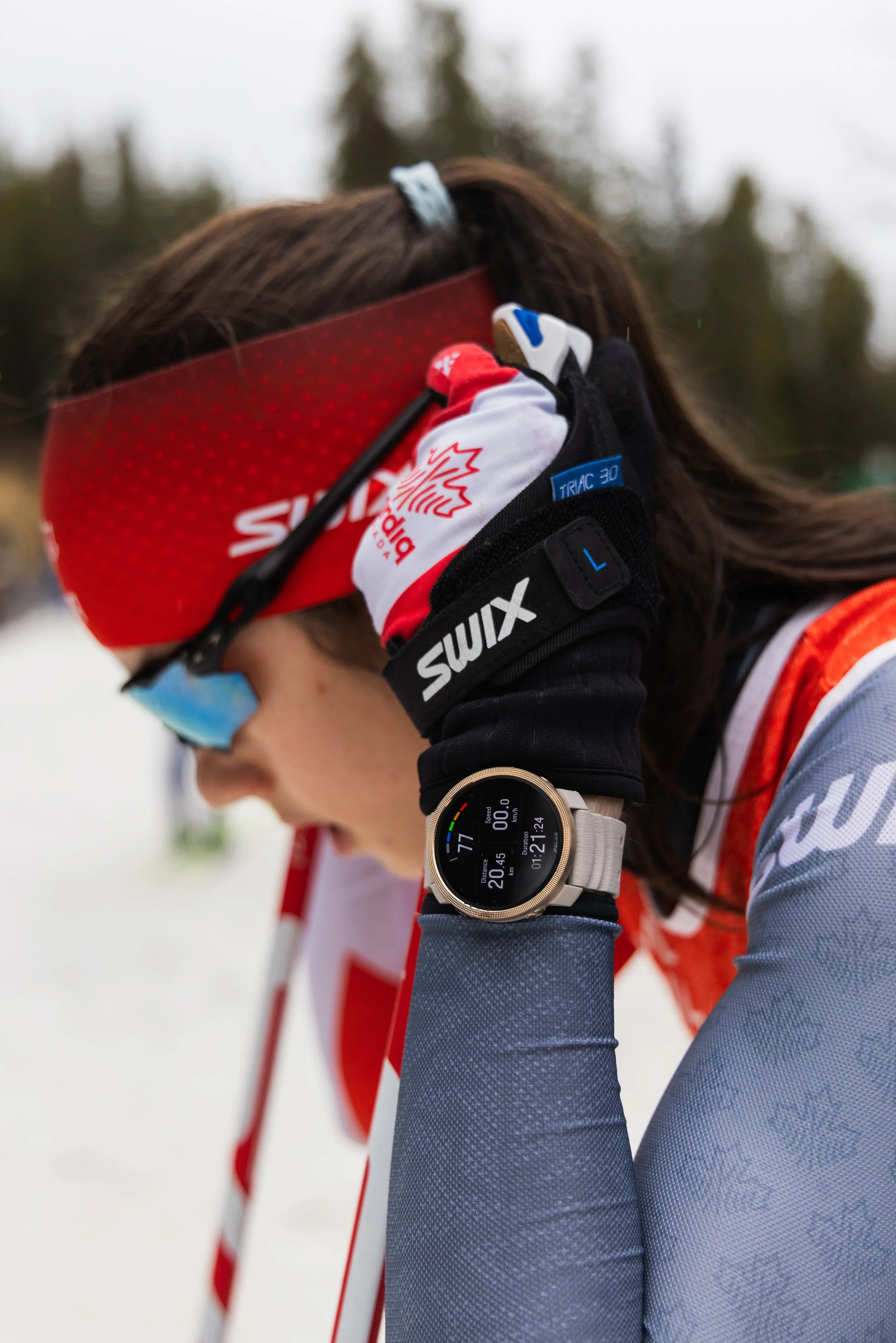 Close-up of an athlete adjusting their ear while competing, wearing a red headband, black gloves, and a sports watch, with a snowy outdoor background.