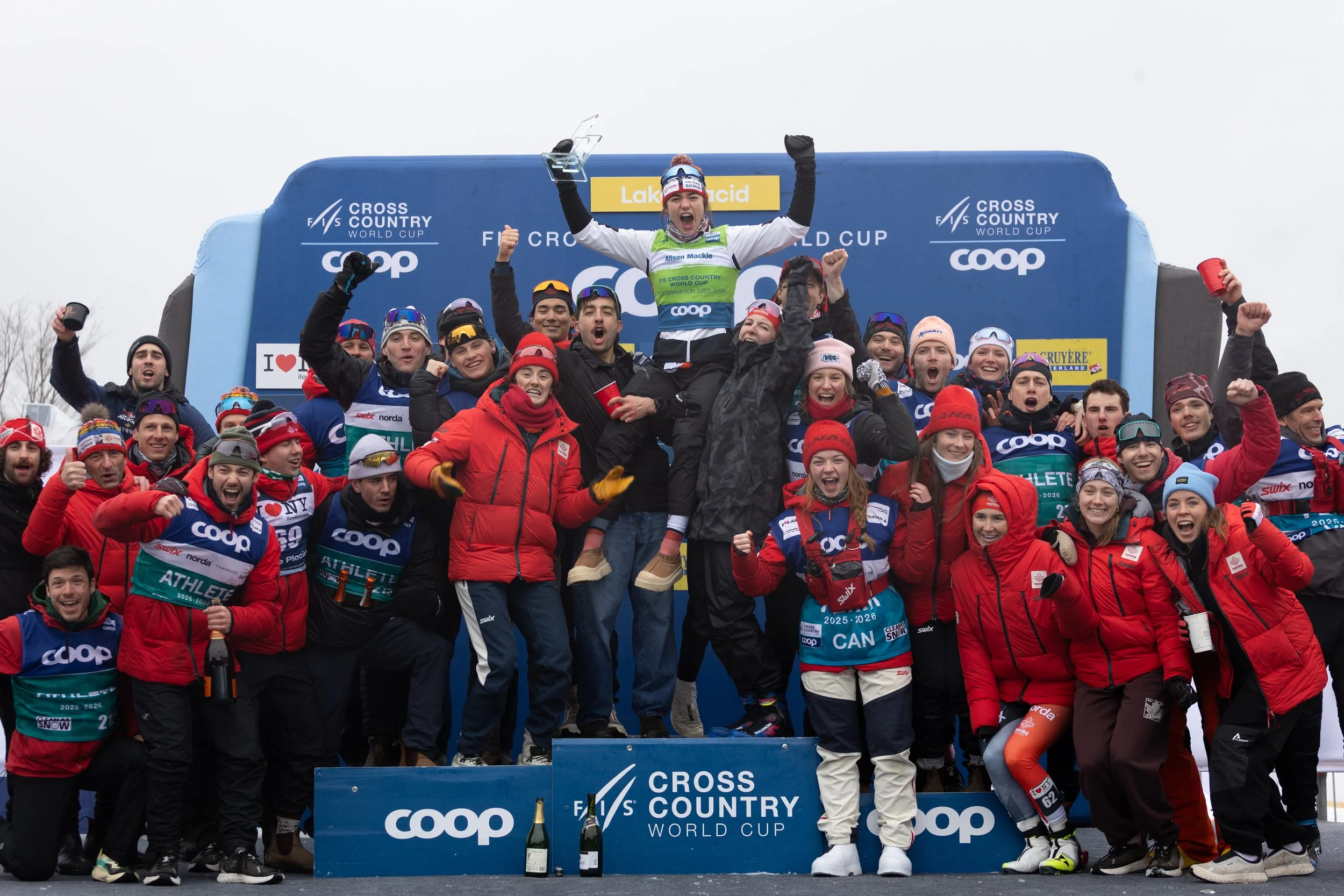 Group of athletes celebrating on a podium at the Cross Country World Cup event, with a woman holding a trophy, in snowy outdoor setting.