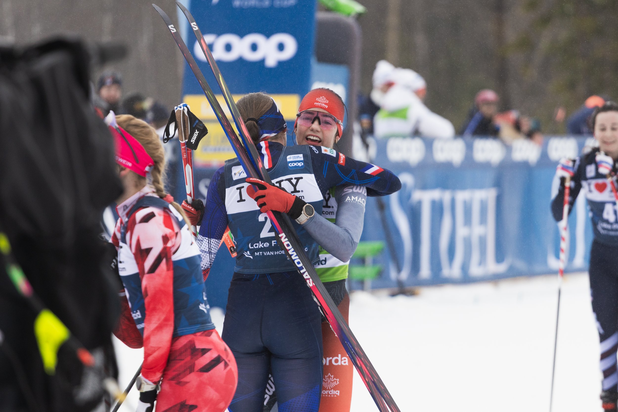 A group of cross-country skiers celebrating at the finish line during a race, with one woman smiling and hugging another, in a snowy outdoor setting with spectators and banners in the background.