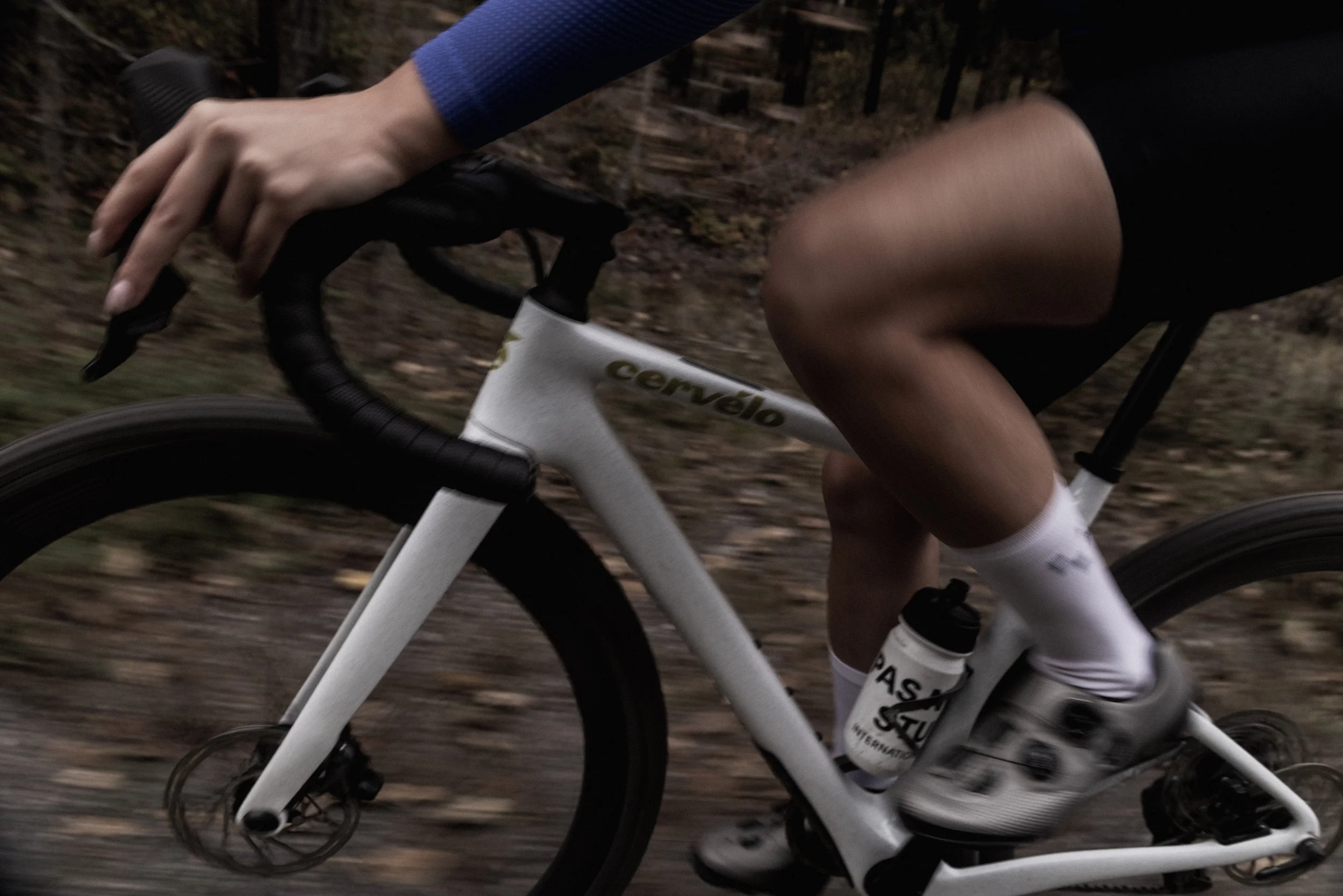 Close-up of a cyclist's hand gripping the handlebars of a white bicycle, with a wooded trail background, showing the cyclist's black shorts, socks, and part of the torso wearing a blue sleeve.