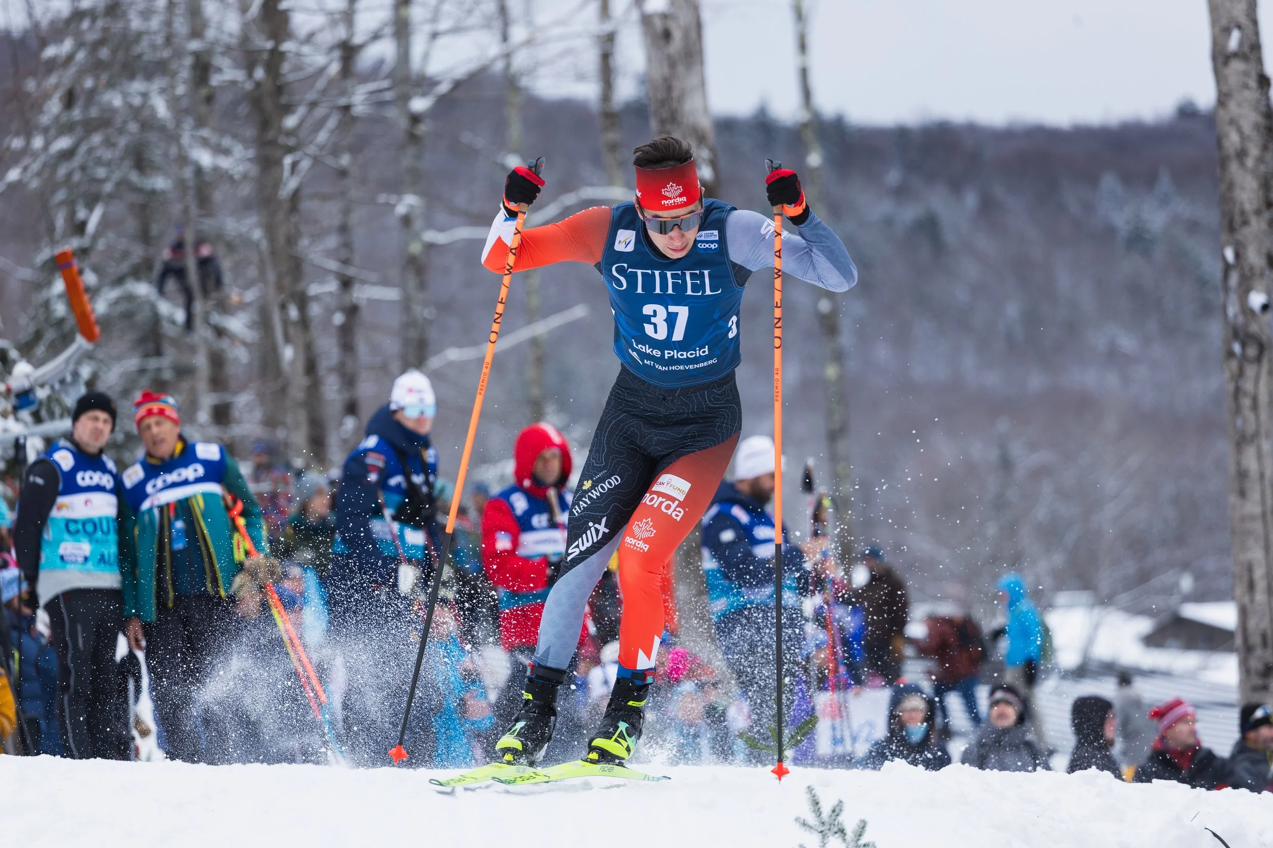A skier in a blue racing suit and red headband skiing downhill in a snowy forest during a winter race.