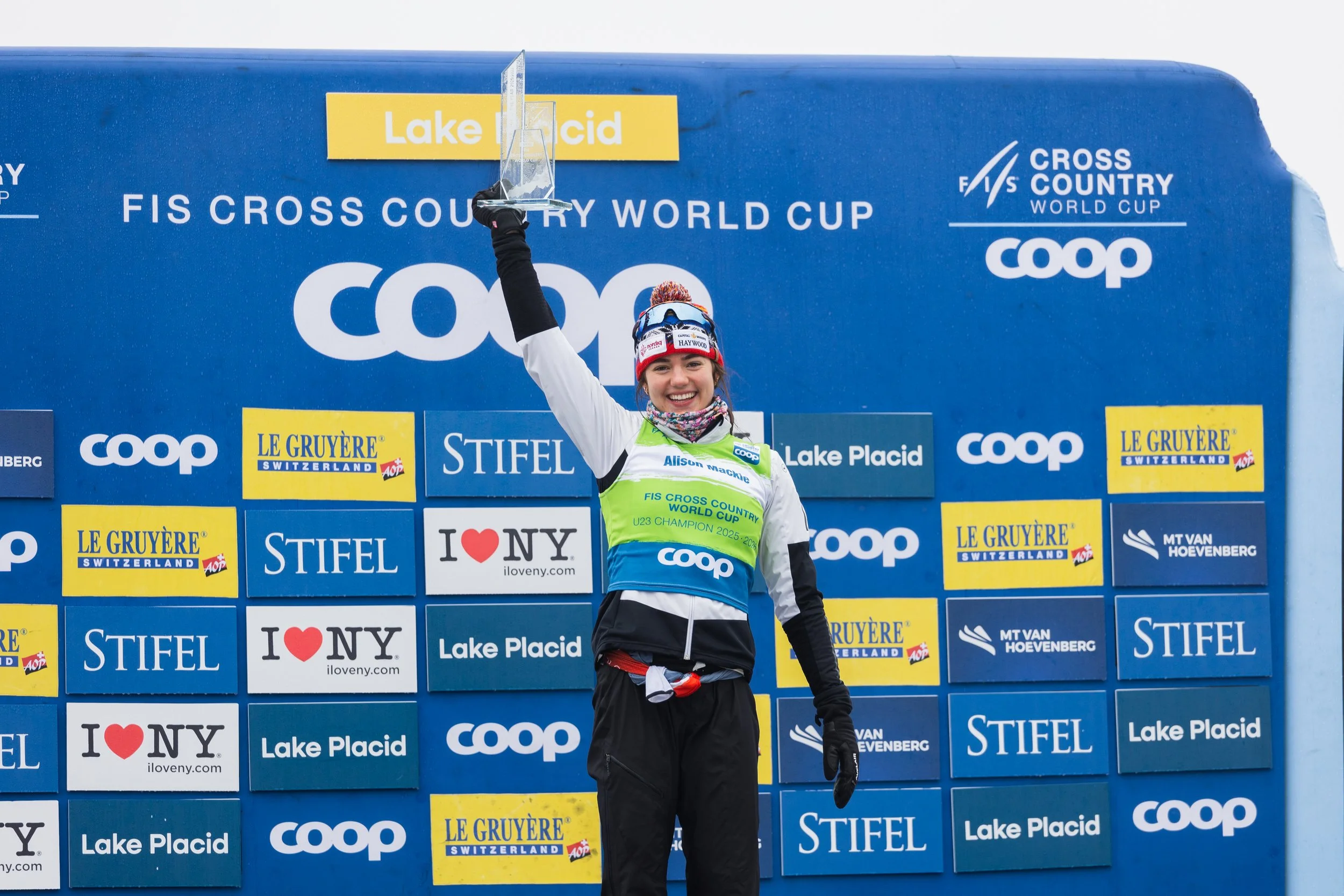 Alison Mackie holding a trophy up high on a podium at the Lake Placid FIS Cross Country World Cup event, smiling and wearing sports gear with sponsor logos.