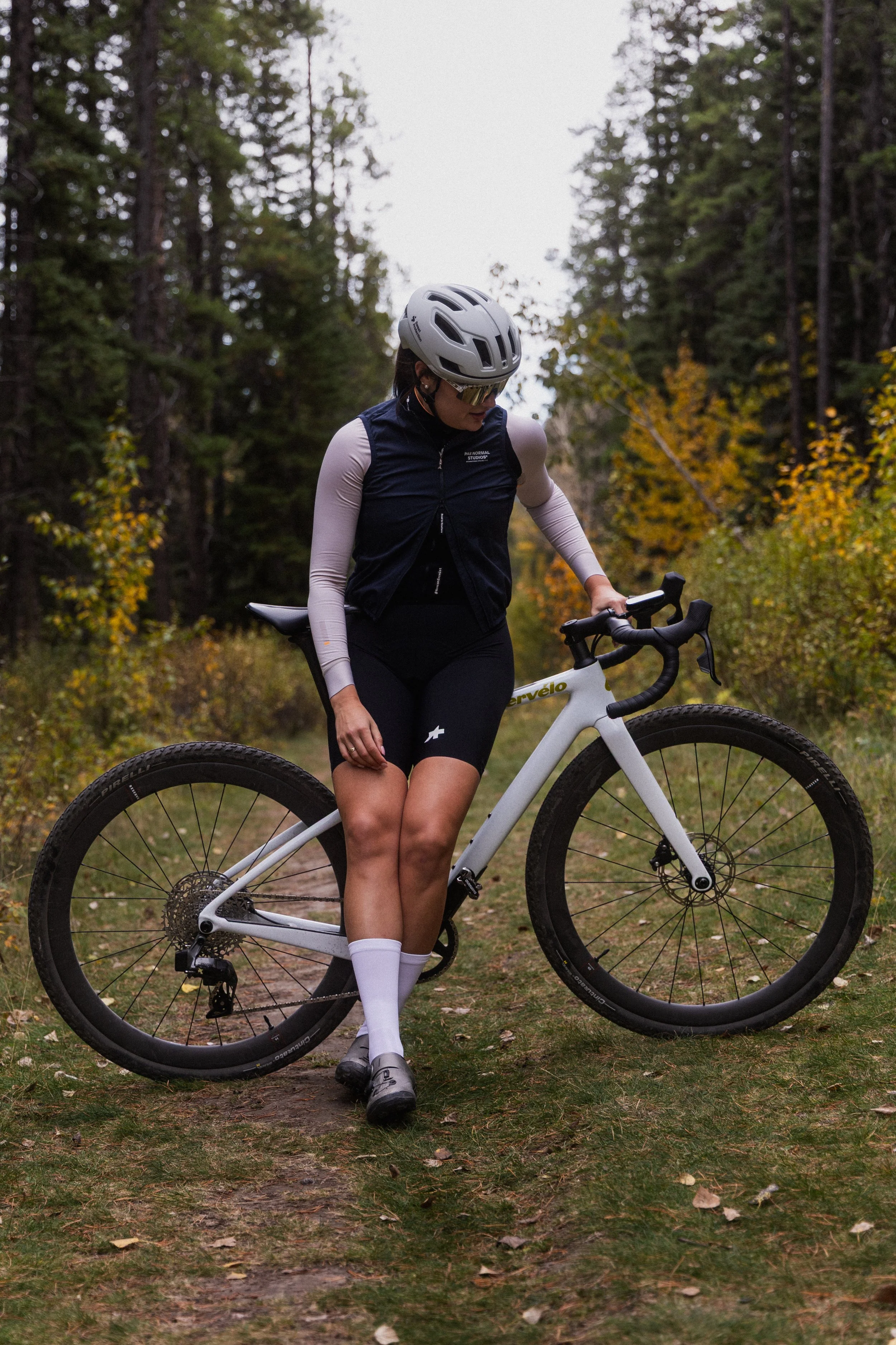 A cyclist wearing a white helmet, sunglasses, a dark blue vest, black shorts, and white socks stands on a dirt trail surrounded by trees with autumn-colored leaves.
