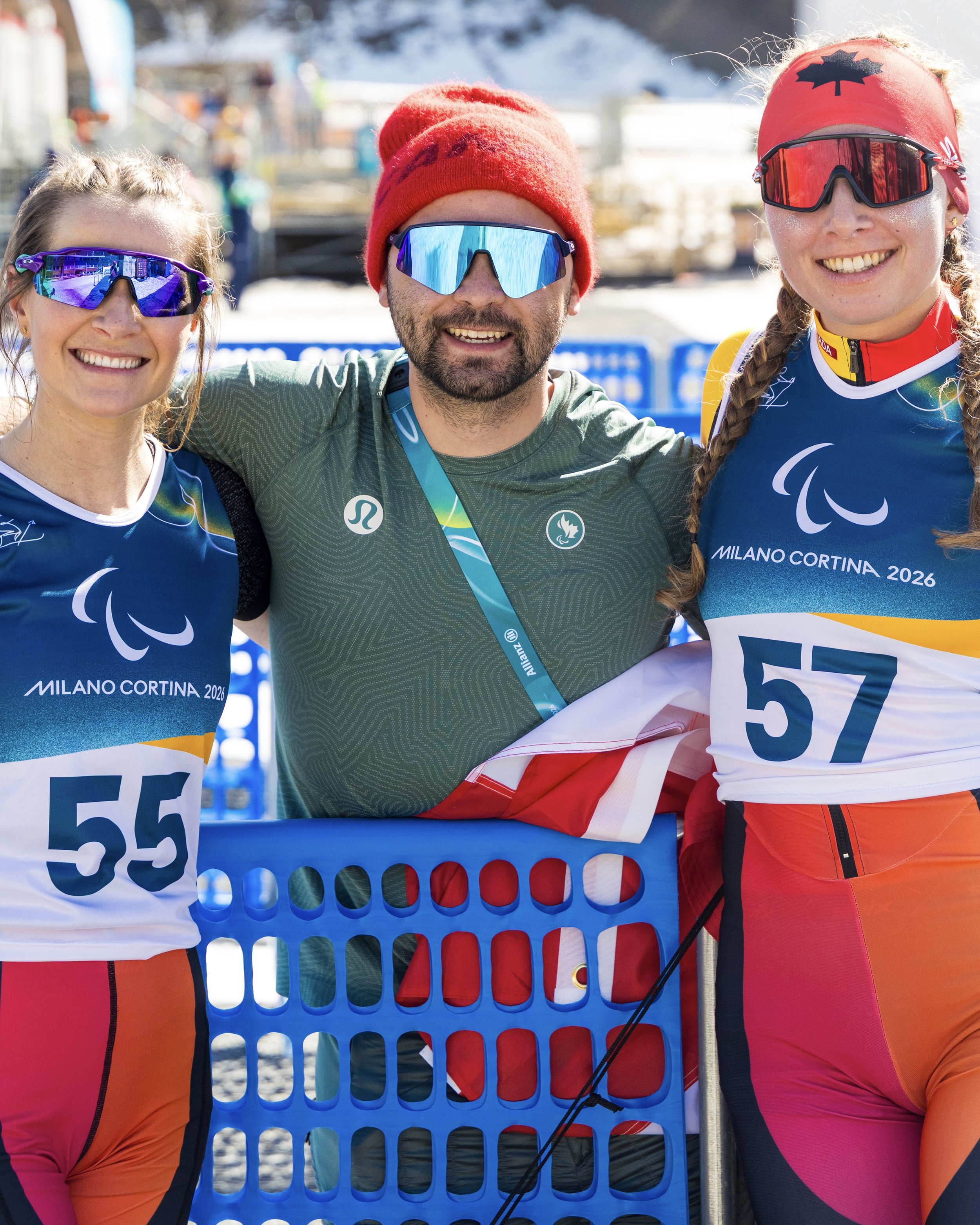 Three athletes in colorful winter sports gear and sunglasses standing close together and smiling at a winter sports event, with snow and blue fencing in the background.