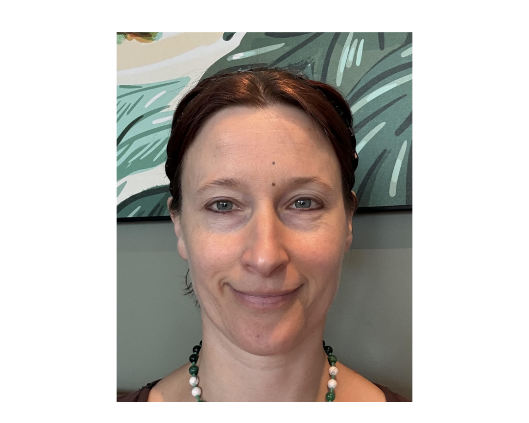 Close-up portrait of a woman with short dark hair and a gentle smile, wearing a beaded necklace against a gray wall and leafy artwork; calm, friendly mood.