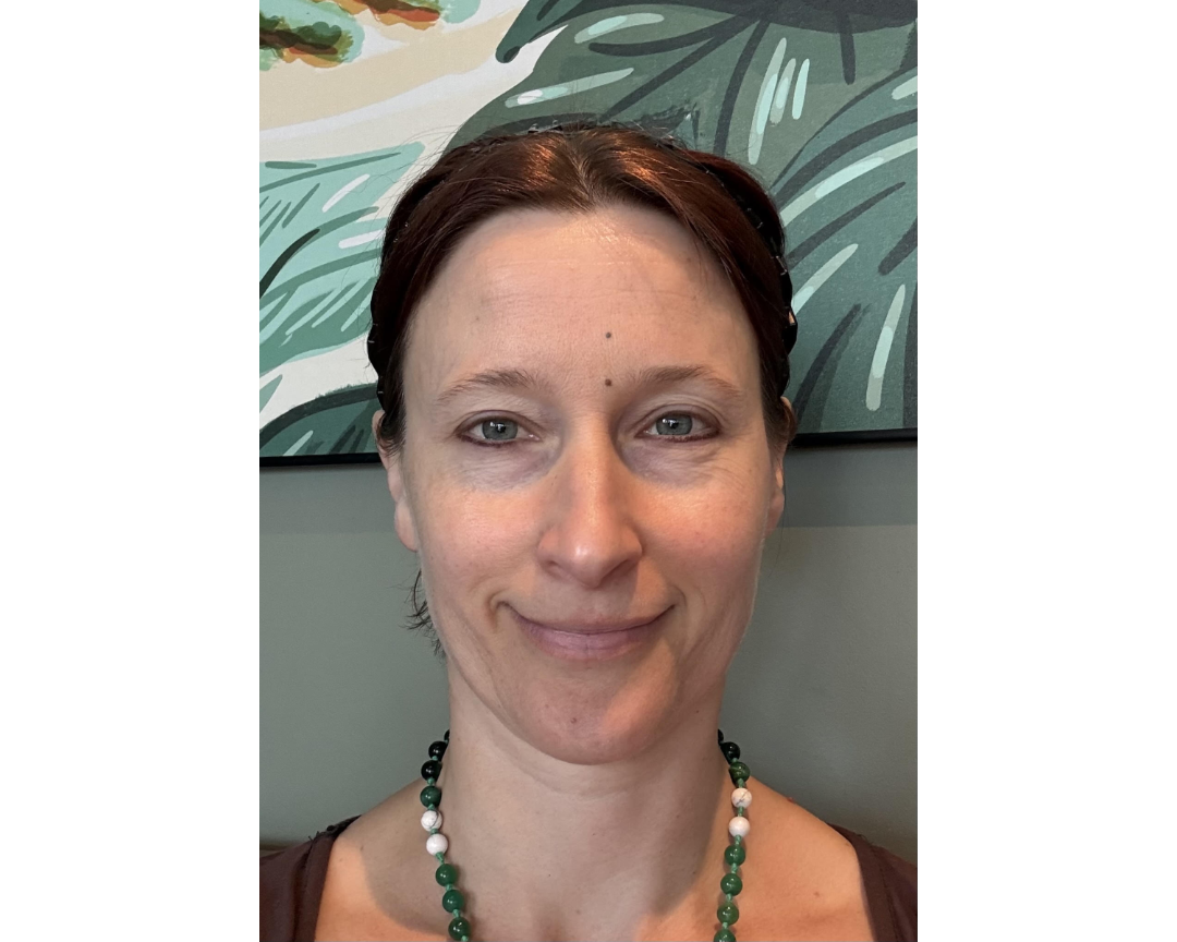 Close-up portrait of a woman with short dark hair and a gentle smile, wearing a beaded necklace against a gray wall and leafy artwork; calm, friendly mood.
