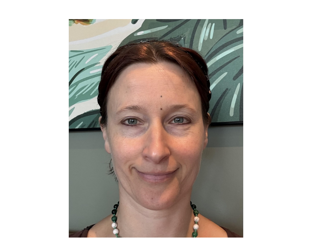 Close-up portrait of a woman with short dark hair and a gentle smile, wearing a beaded necklace against a gray wall and leafy artwork; calm, friendly mood.