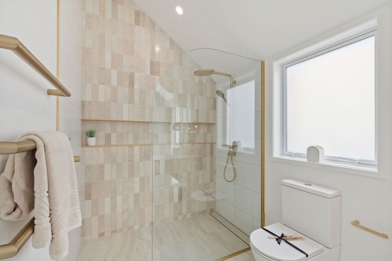 Modern bathroom with a glass-enclosed shower, beige tiled walls, a white toilet with a decorative black ribbon, a frosted window, and gold accents.