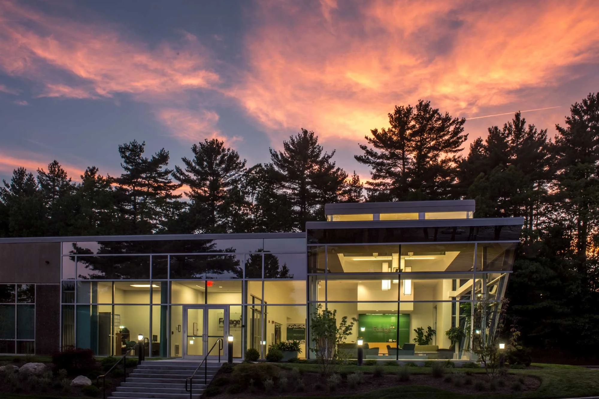 Modern glass building with interior lighting, trees in front, and a colorful sunset sky with pink and orange clouds overhead.