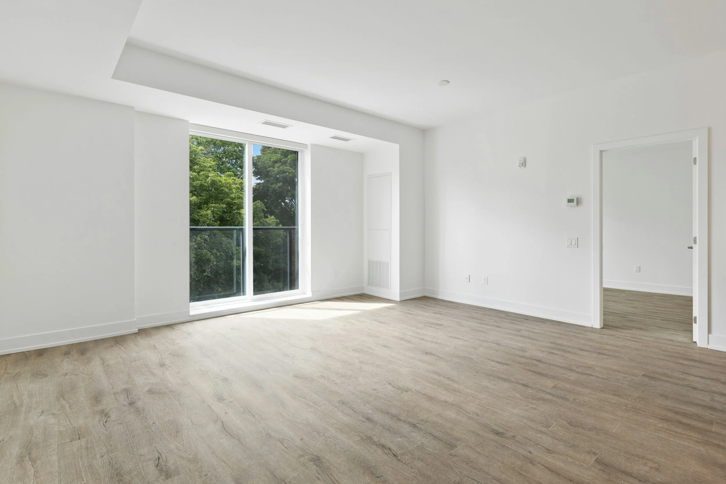 Empty room with large window showing trees outside, white walls, and light wood flooring.