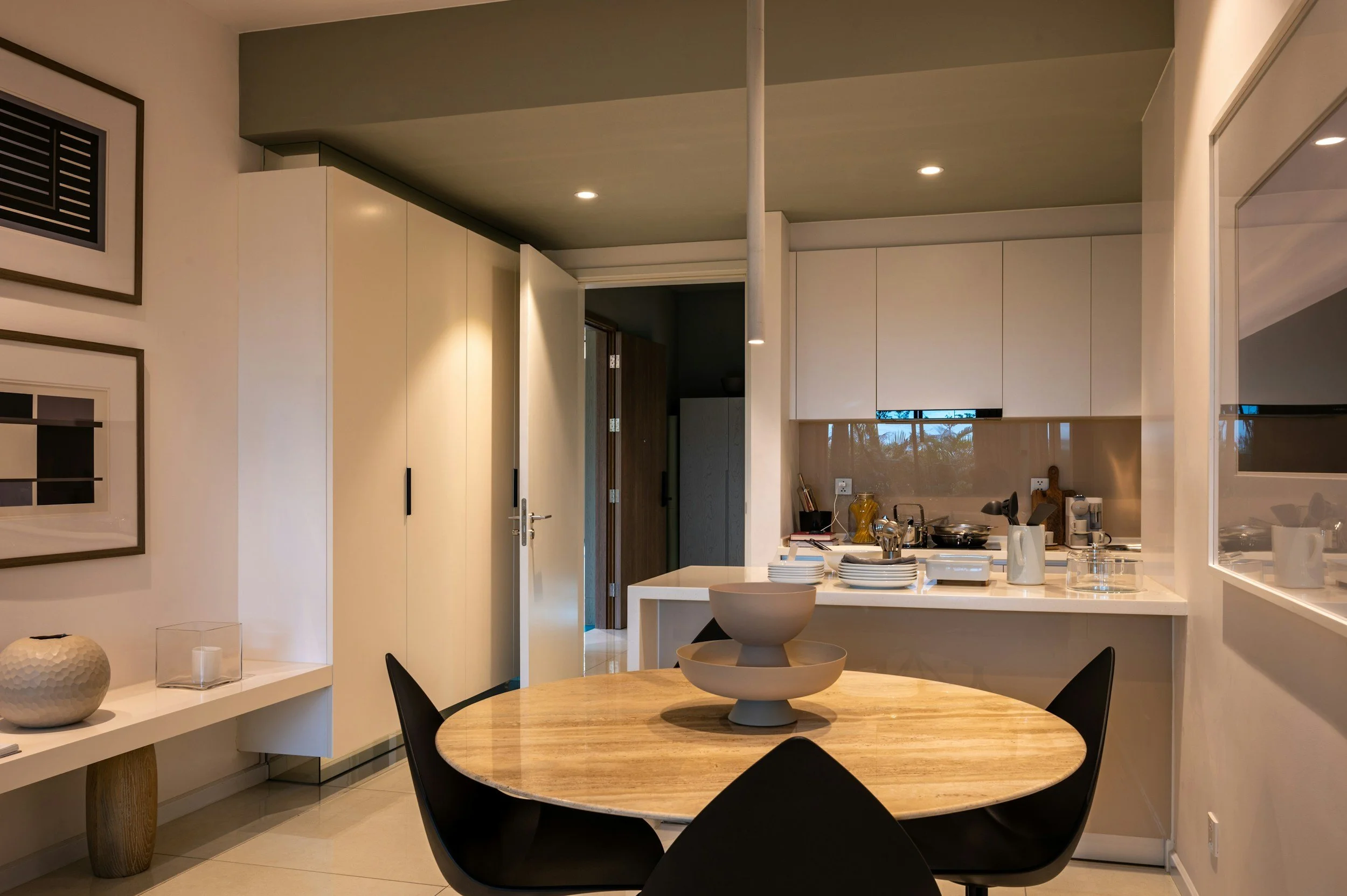 A modern kitchen with white cabinets, a wooden dining table with black chairs, and a round wooden centerpiece. There are some plates, cutlery, and kitchen utensils on the counter, with a doorway leading to another room. Decorative framed artwork hangs on the wall.