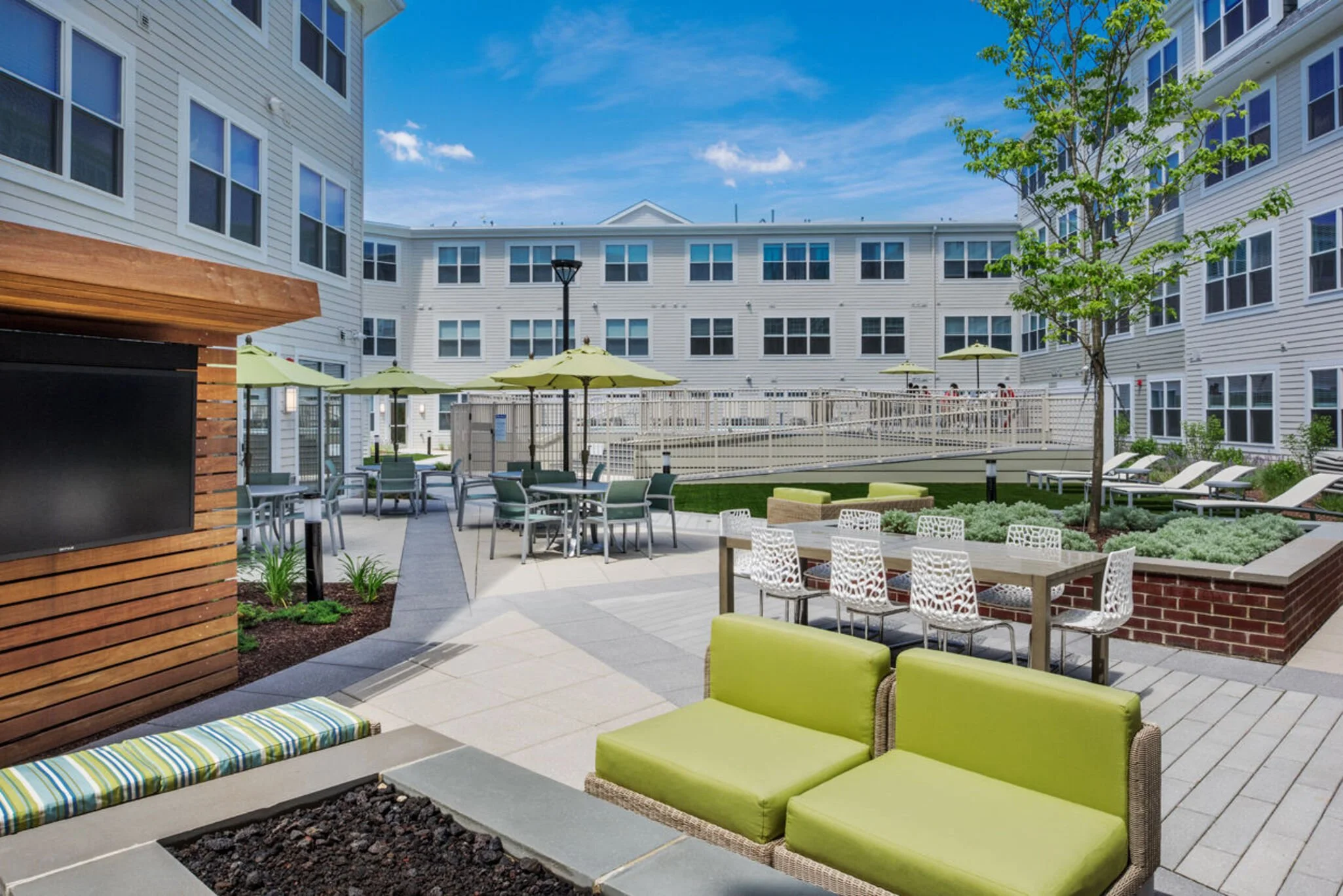 Outdoor courtyard with green cushioned lounge chairs, patio tables, yellow umbrellas, and surrounding white multi-story buildings under a blue sky.