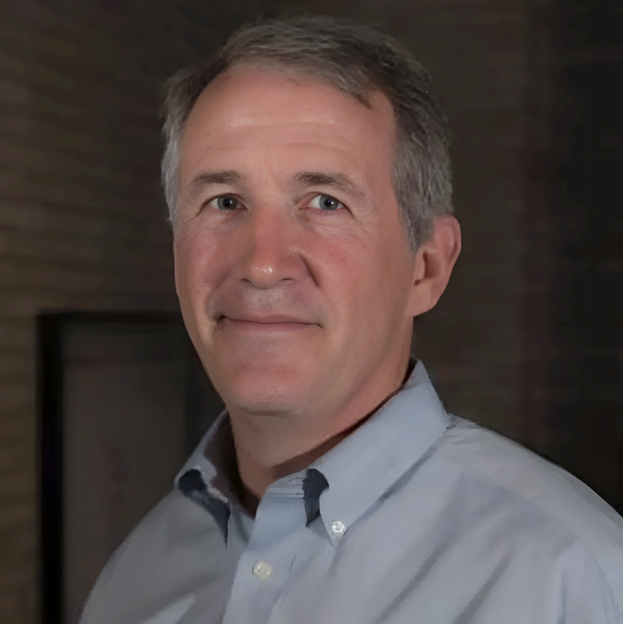 Headshot of a middle-aged man with short, gray hair, wearing a light gray collared shirt, standing in front of a dark, textured wall.