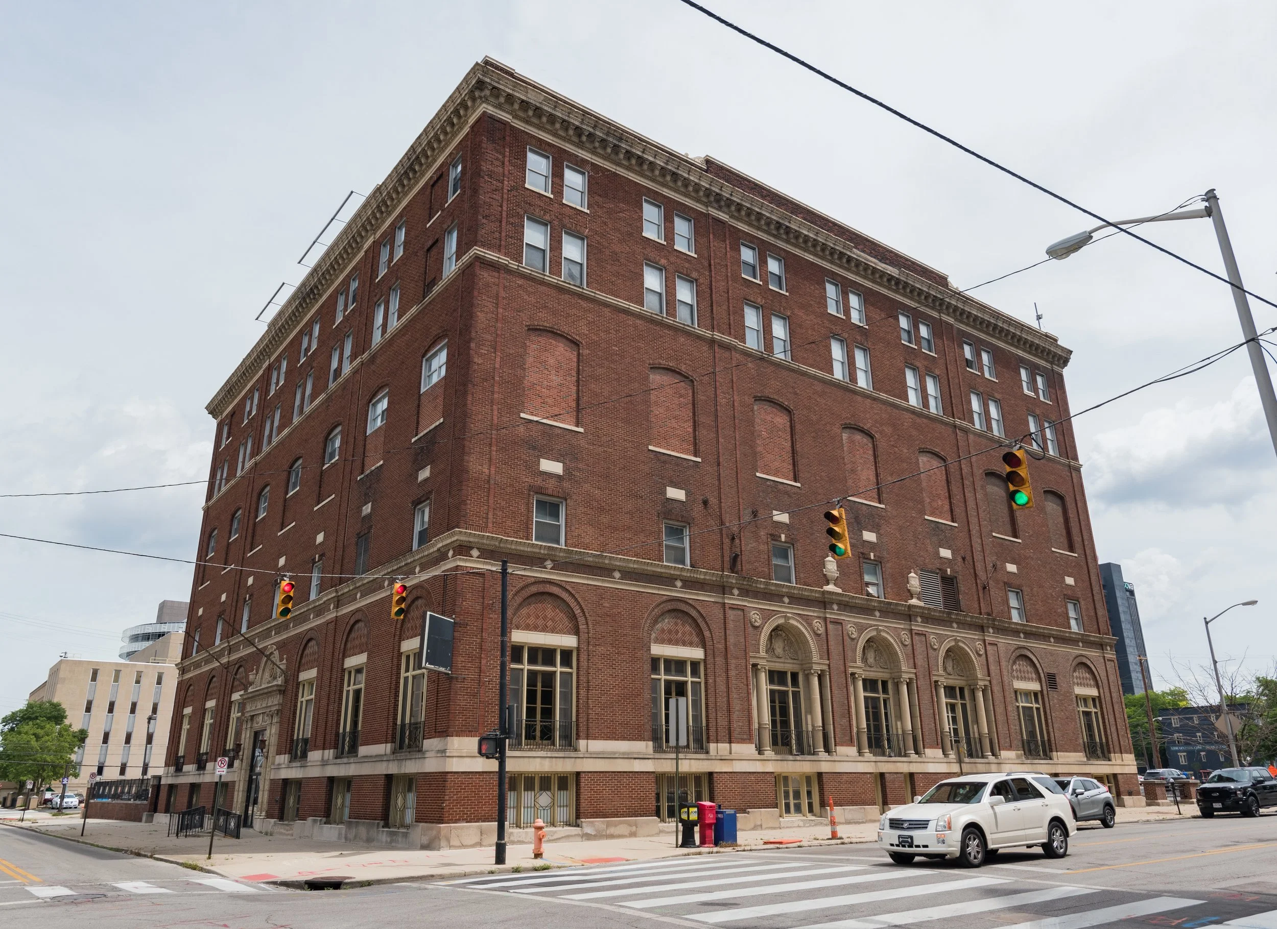 An exterior view of a multi-story brick apartment building with balconies and large windows. There is a grassy area with small shrubs in front of the building and a large tree on the left side. The sky is clear with a few clouds, and sunlight is shining from the left side of the image.