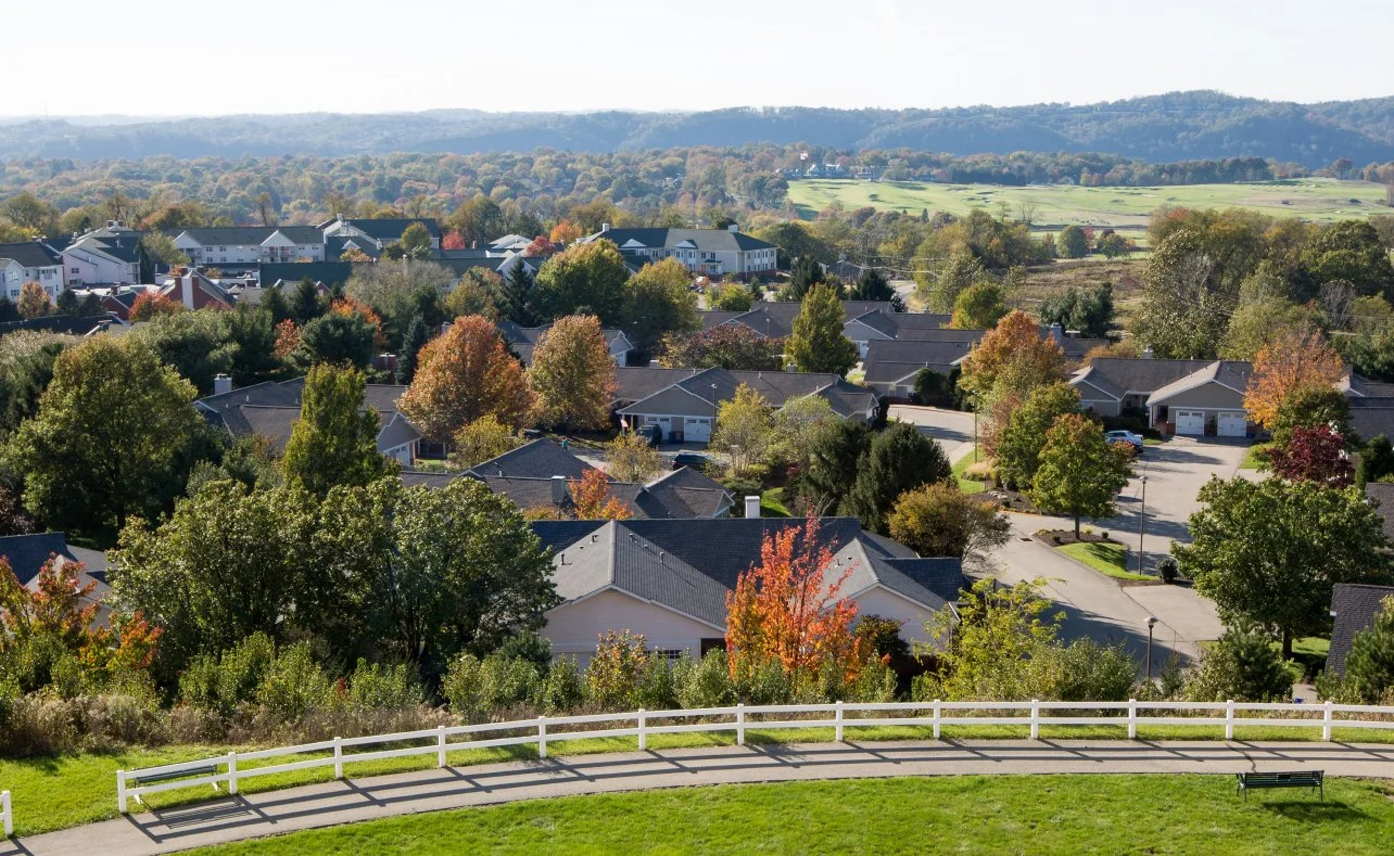 A suburban neighborhood with houses, trees with fall foliage, and a hilly landscape in the background.
