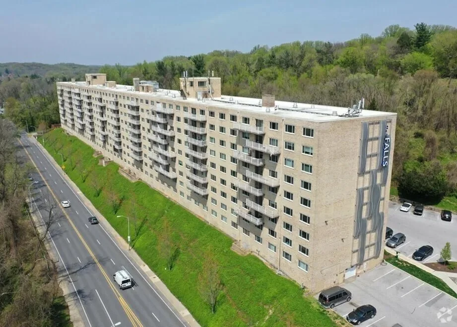 A modern white apartment building surrounded by lush green lawns and various flowering trees under a clear blue sky.